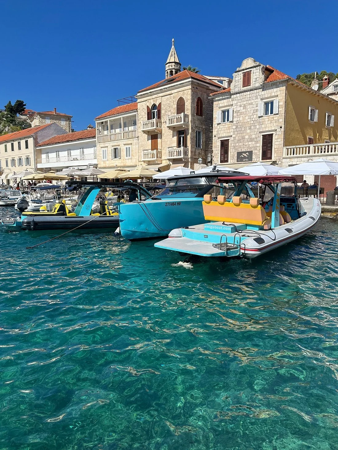 Boats docked on clear turquoise water in front of historic stone buildings with red-tiled roofs and white shutters, under a bright blue sky.
