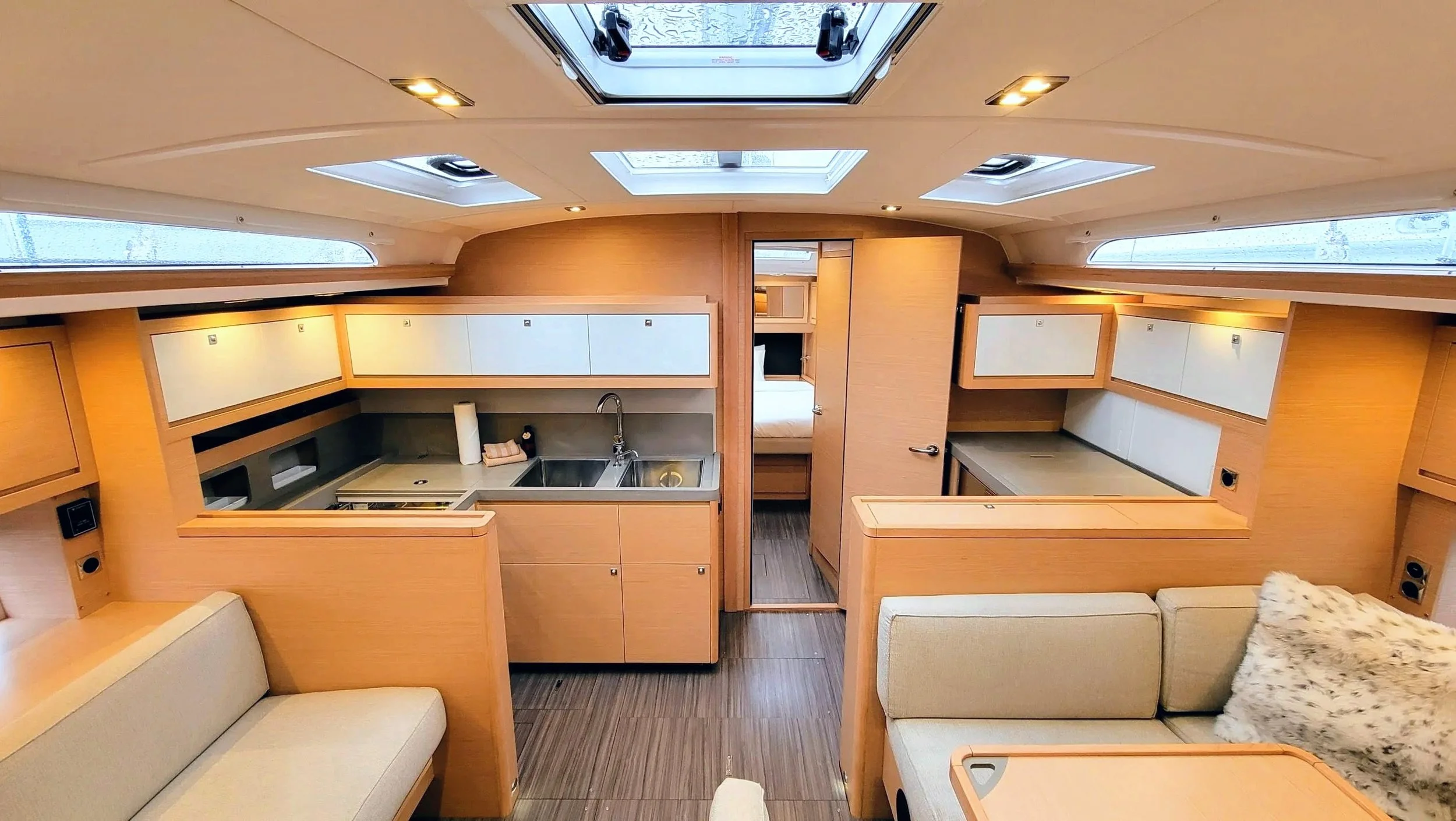 Interior of a boat or yacht with a small kitchen area, sofa, skylights, and sleeping quarters visible through an open door.