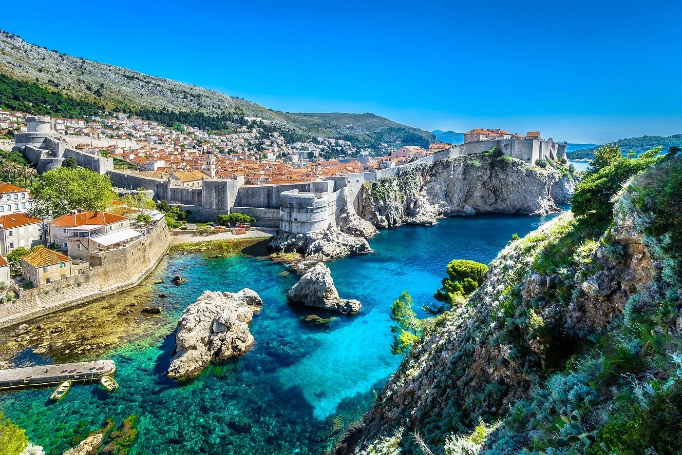 Historical city with stone fortress walls along the coast, lush greenery, and clear turquoise water.