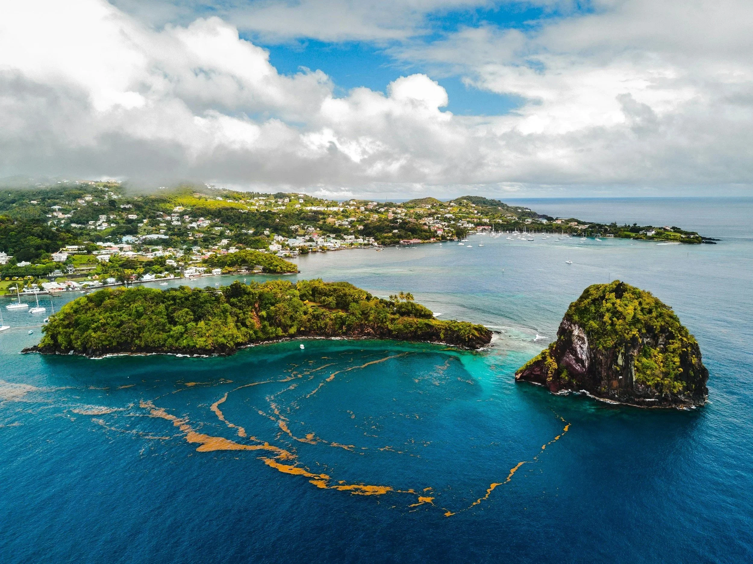 An aerial view of a coastal area with lush green islands, a bay with sailboats, and a residential area on hilly terrain under partly cloudy skies.