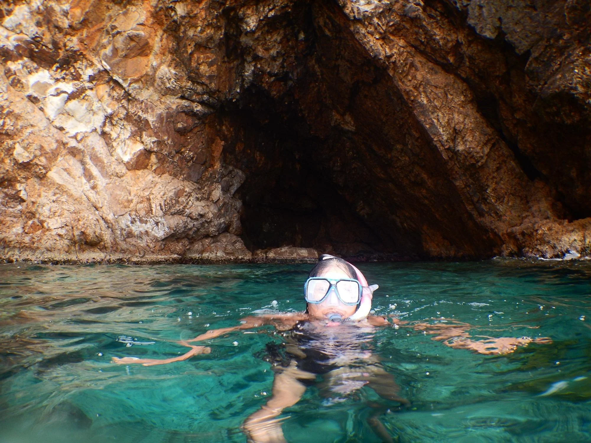 A person wearing snorkeling gear swimming near a rocky cave opening in clear water.