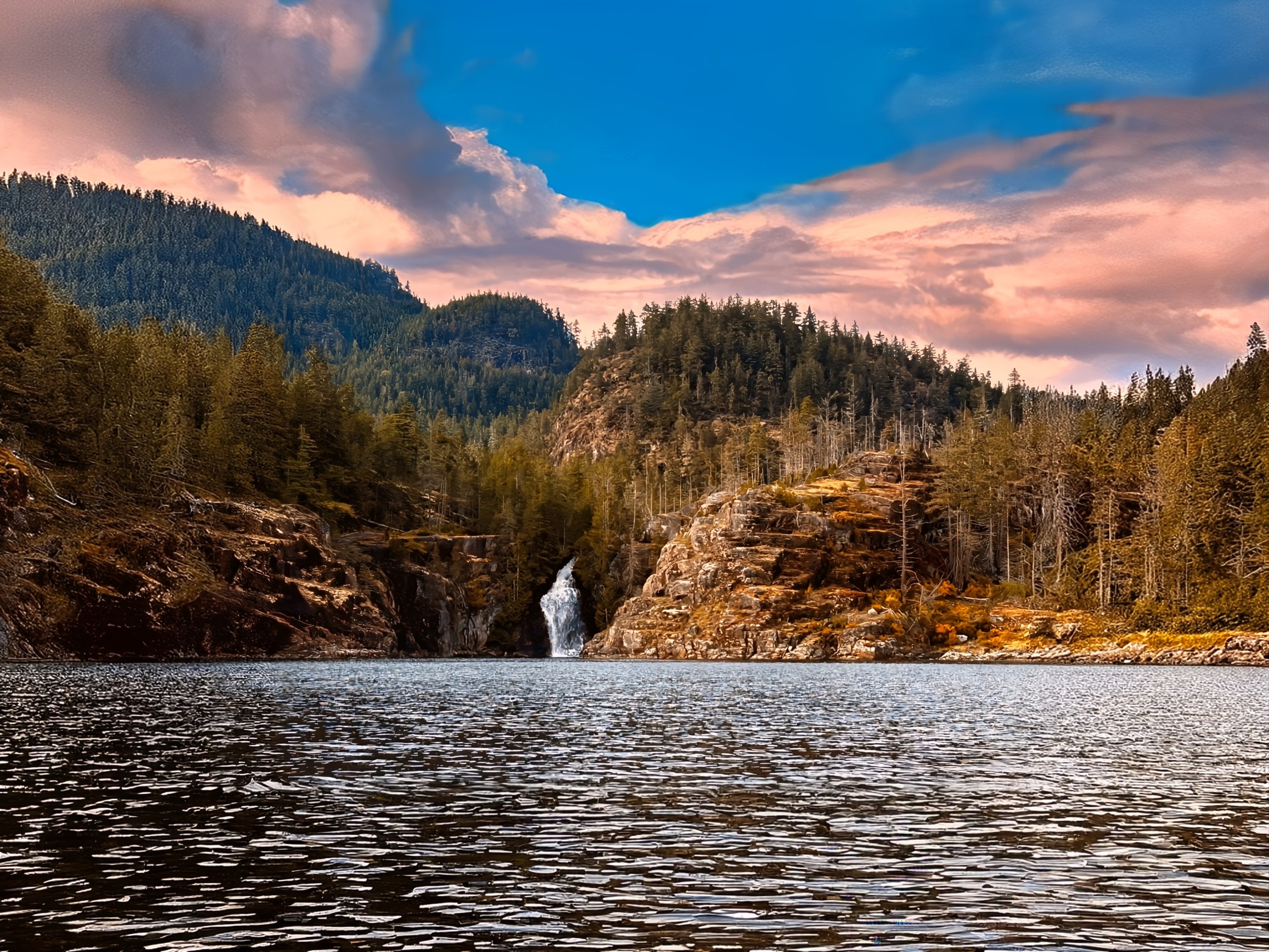 A scenic landscape featuring a lake with rippling water, surrounded by forested hills and mountains, with a waterfall cascading down rocks into the lake, under a colorful sky with clouds.