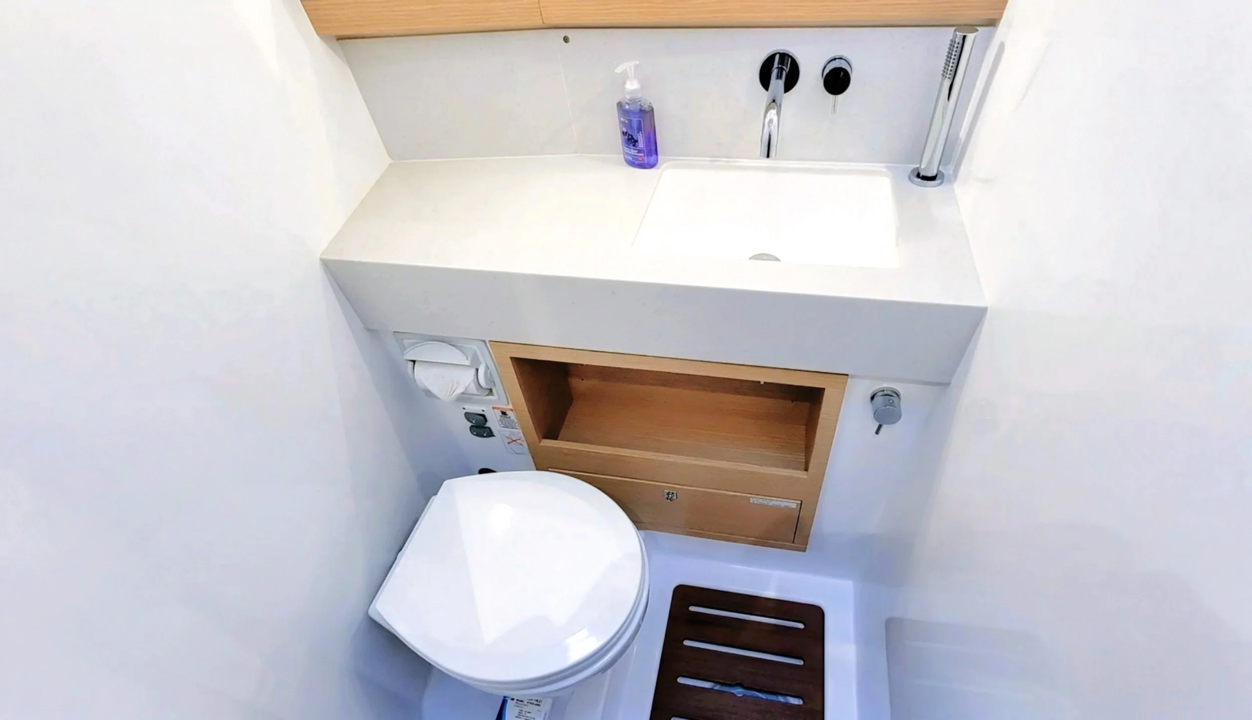 A small bathroom with a white countertop sink, a purple soap dispenser, a mounted faucet, a small wooden cabinet below the sink, a white toilet with a paper holder, a black bath mat, and a soap or lotion dispenser on the wall.