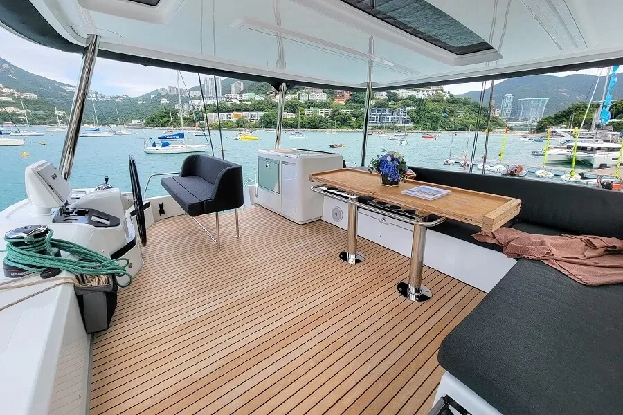 Interior of a yacht with a view of the harbor, showing a boat's control panel, a black chair, a small white kitchenette, a wooden table with a blue flower arrangement and papers, and a seating area with a brown cloth, during daytime.