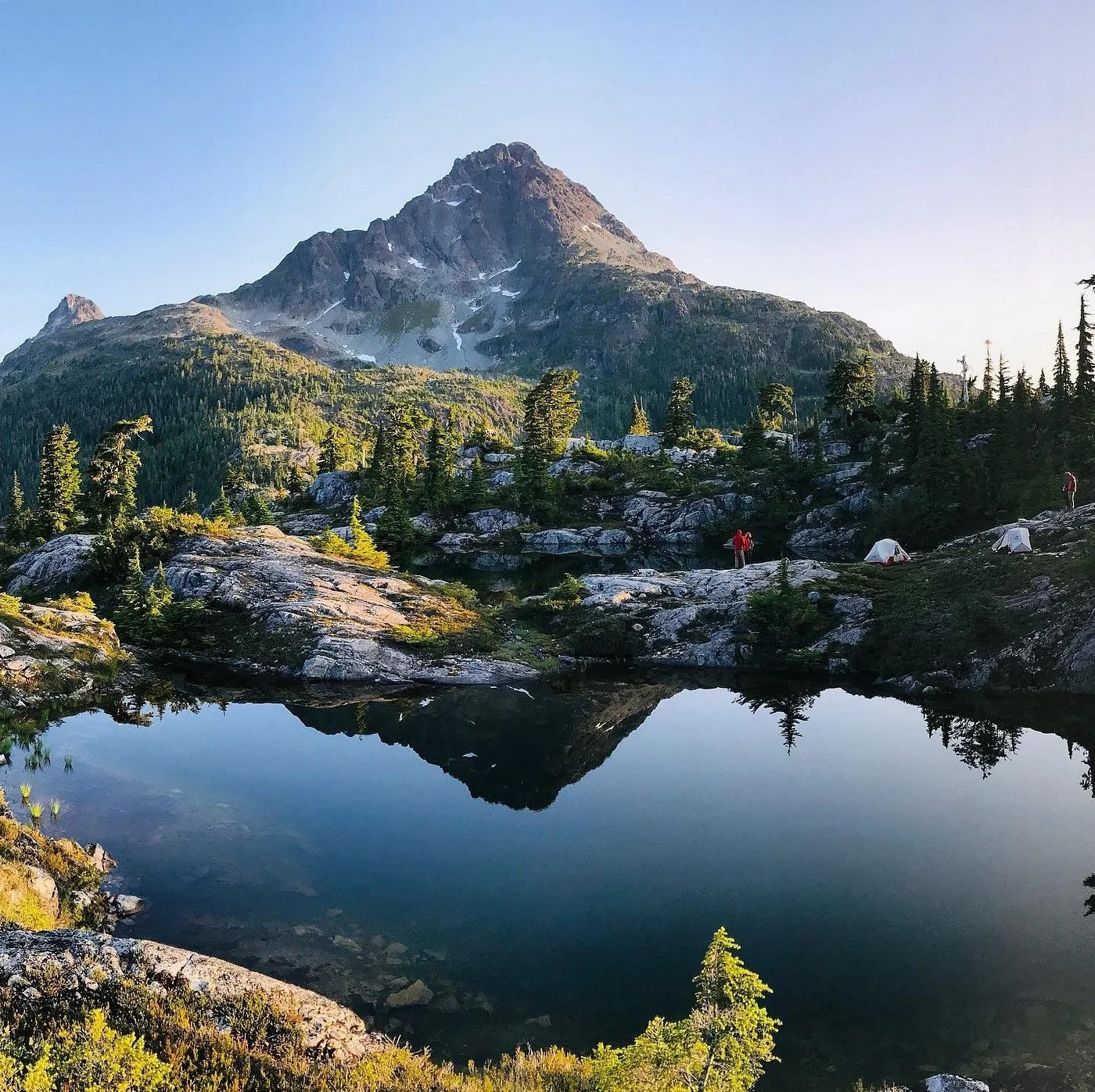 A mountain landscape with a reflection in a lake, trees, and campers on rocks.