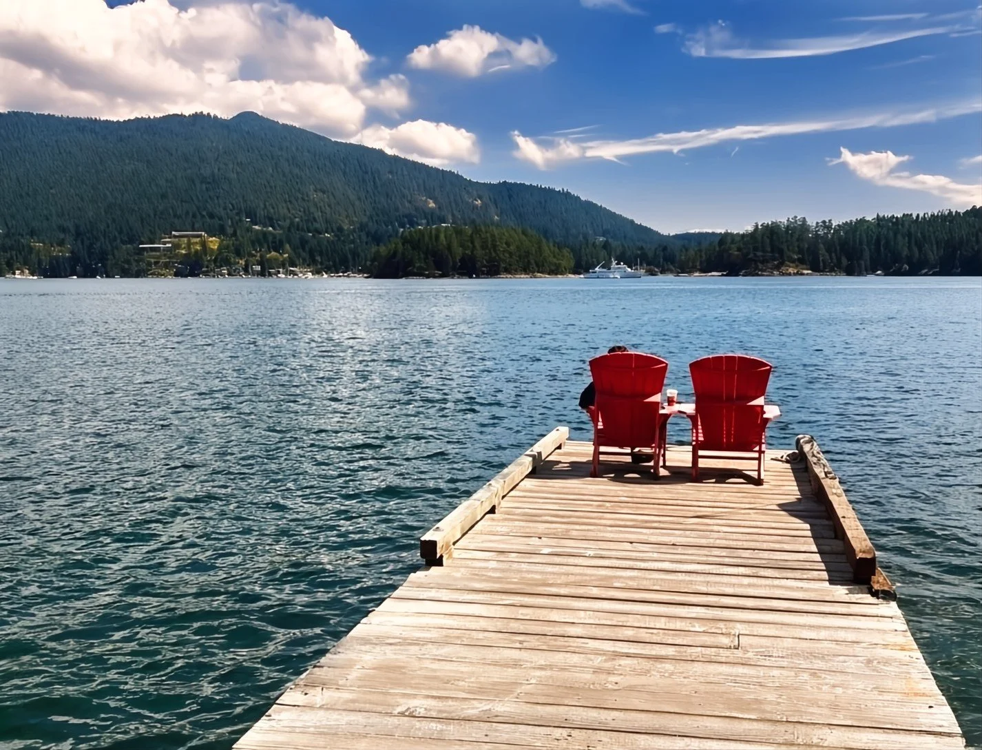 A wooden dock extends over a body of water with two red chairs on it, facing the water. In the background, there are forested hills and a partly cloudy sky.