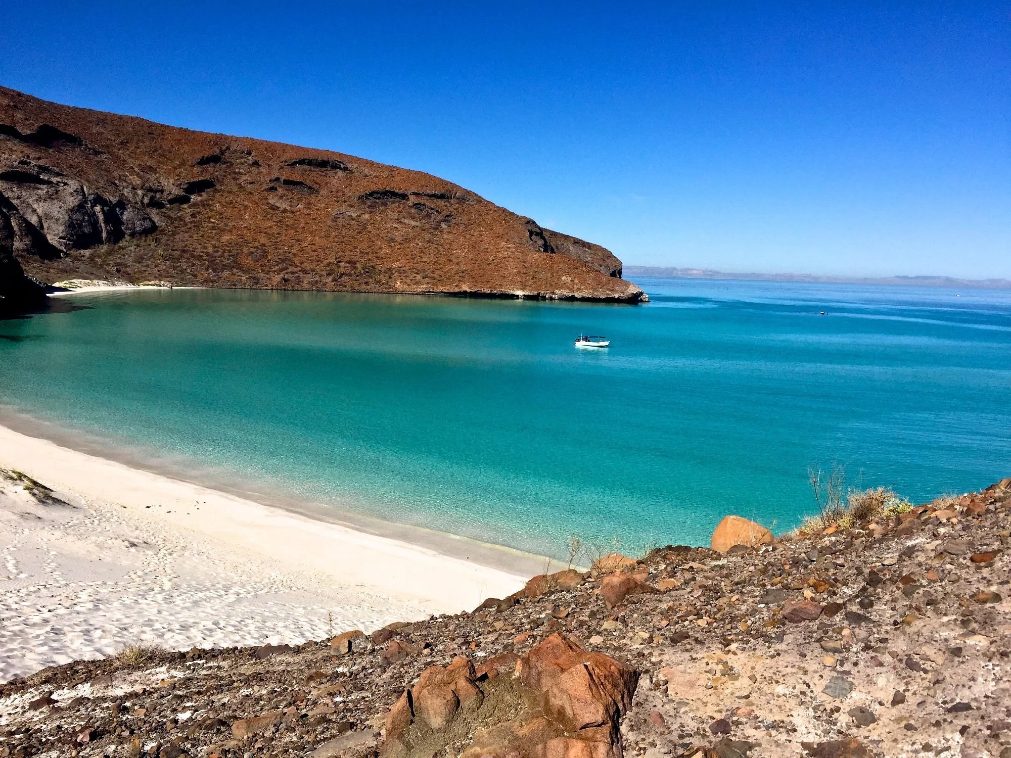 A secluded beach with white sand, turquoise water, a boat, and brown rocky hills under a clear blue sky.