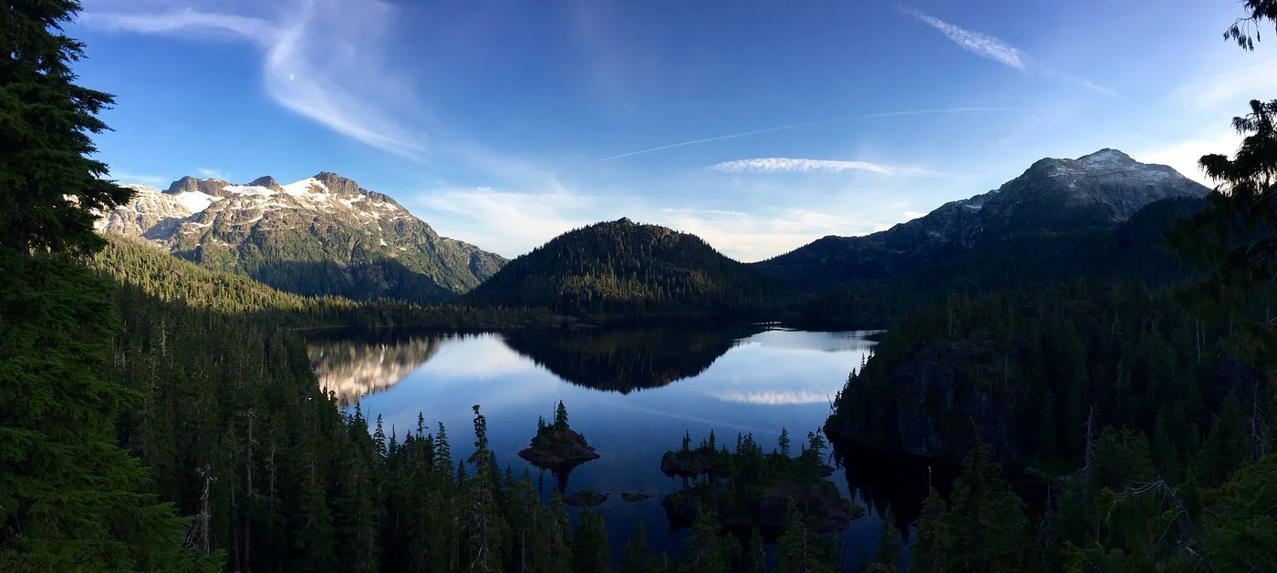 A scenic view of a calm mountain lake surrounded by dense evergreen forests, with snow-capped peaks in the background under a clear blue sky.