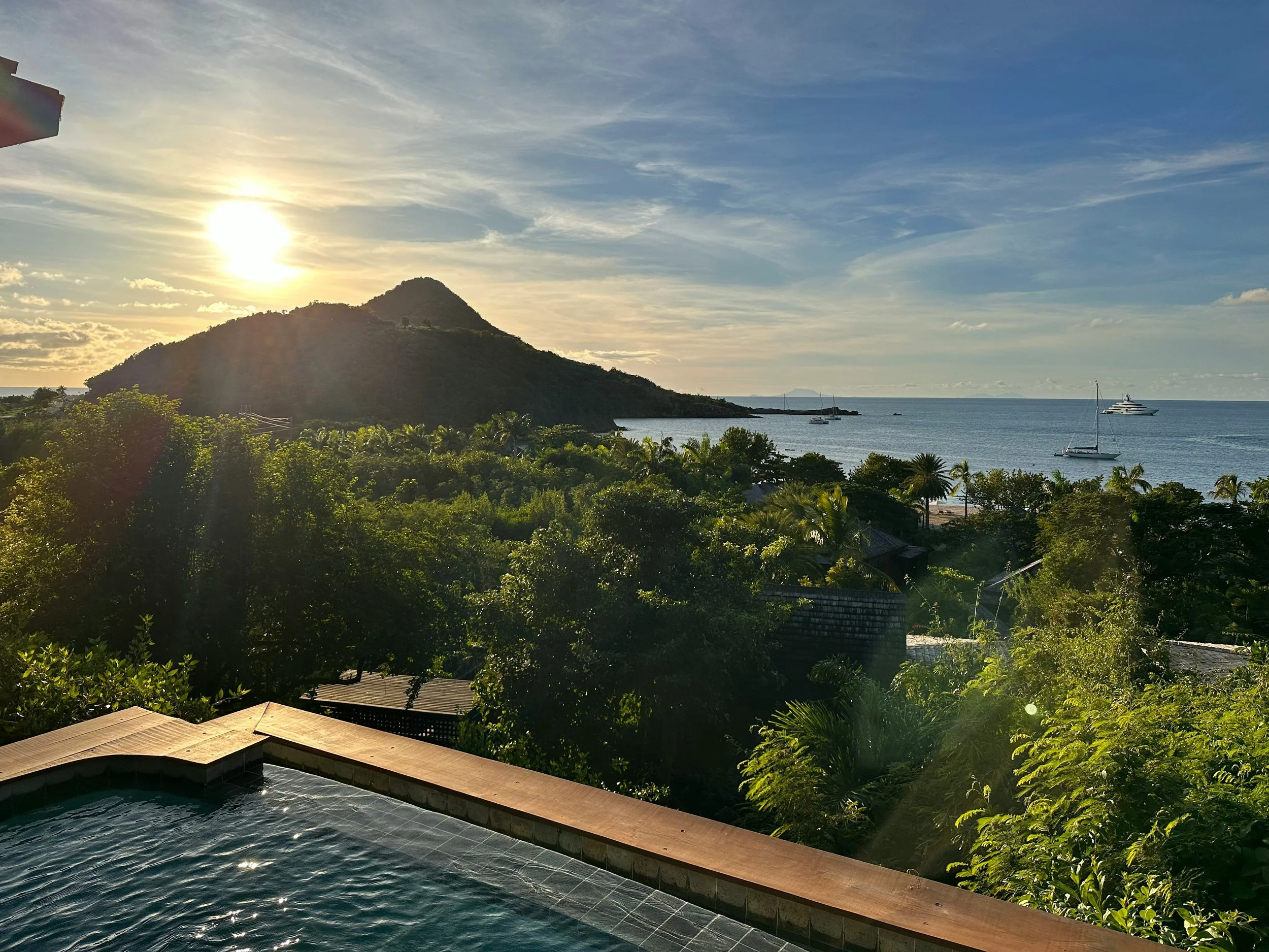 Sunset view over a lush green tropical landscape with mountains, trees, and the ocean, with sailboats and a yacht on the water, and a swimming pool in the foreground.