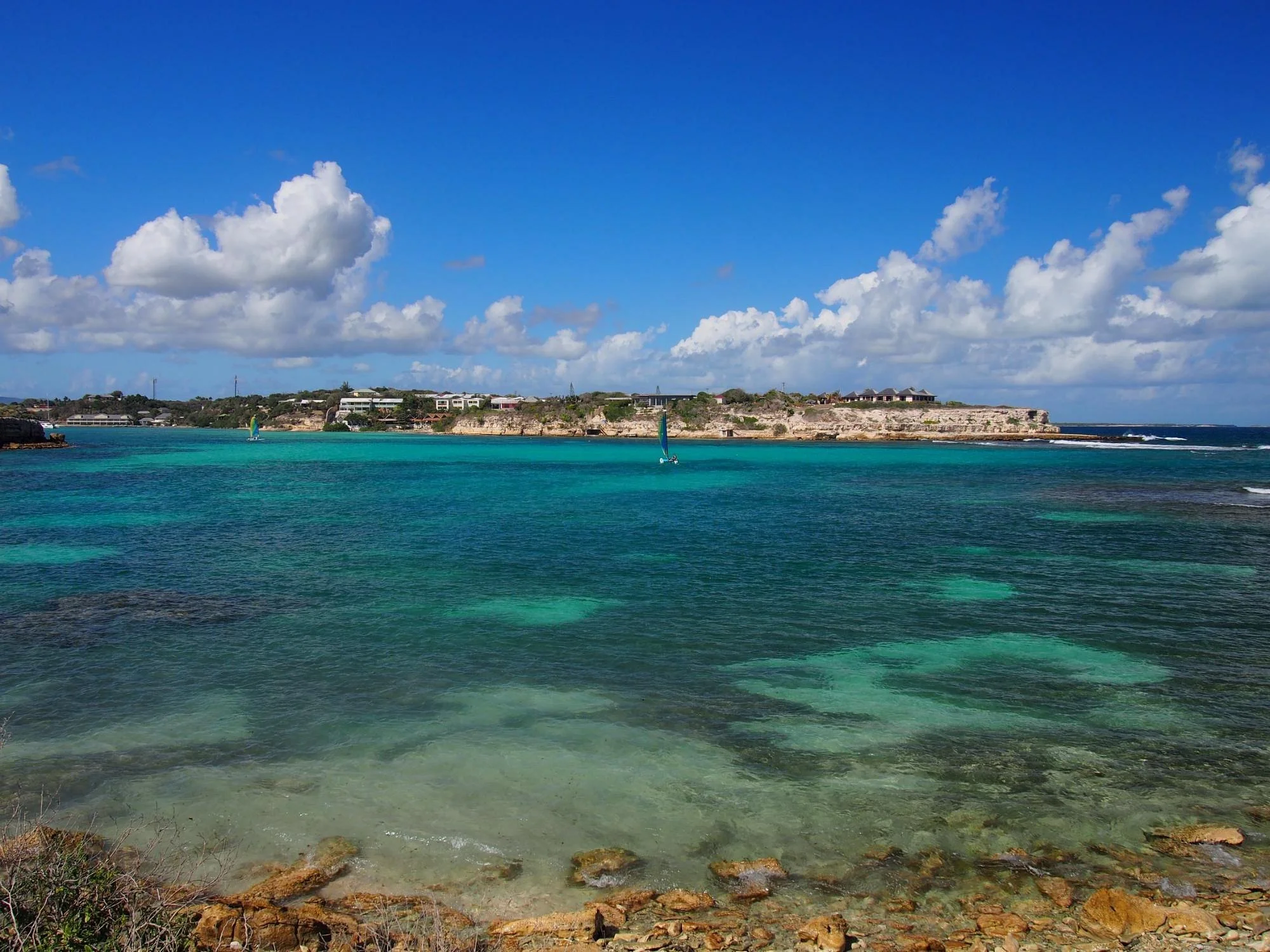 Clear blue ocean with a sandy shoreline in the foreground, a sailboat in the distance, and a rocky cliff with hotels and houses on top under a partly cloudy sky.
