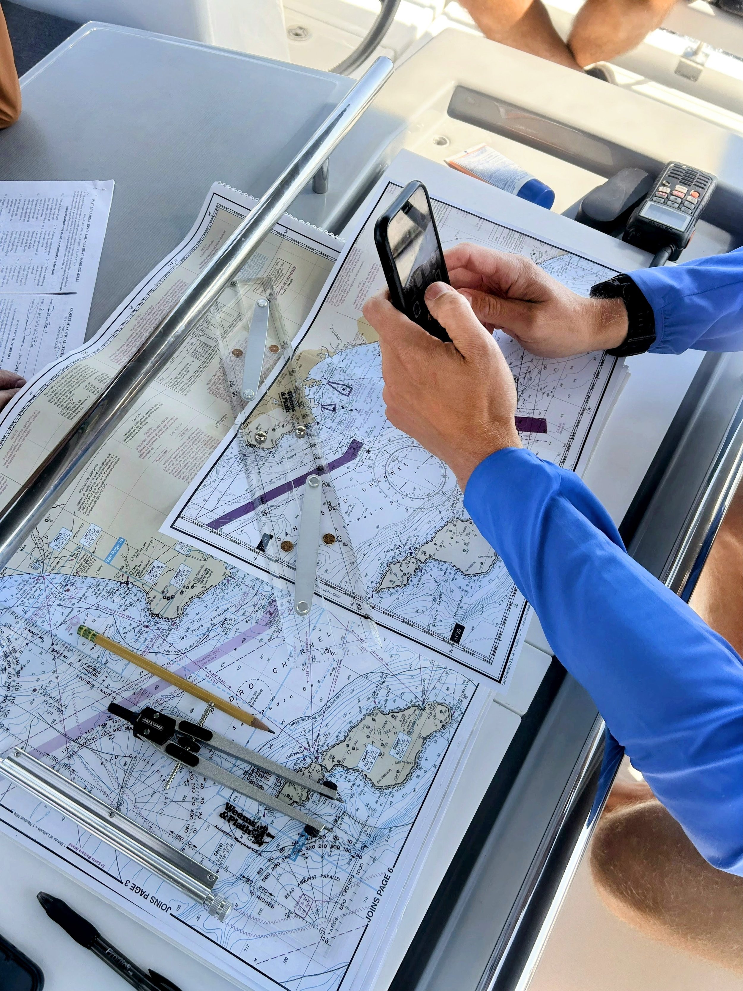 Person in a blue jacket using a smartphone on a marine navigation chart with navigational tools, papers, and a radio on a boat navigation console.