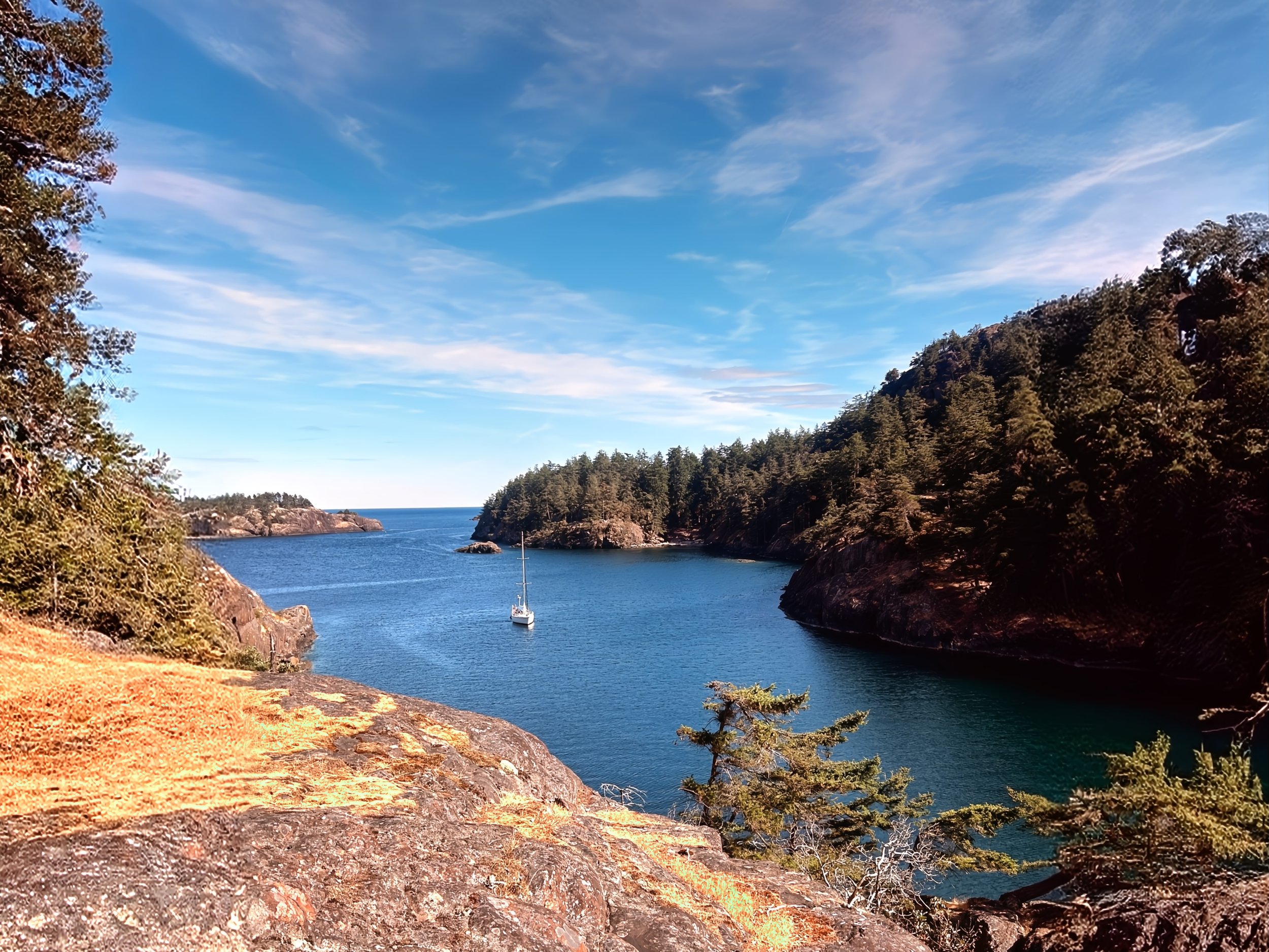 A scenic view of a calm blue bay with a sailboat, surrounded by rocky cliffs and dense green trees under a partly cloudy sky.