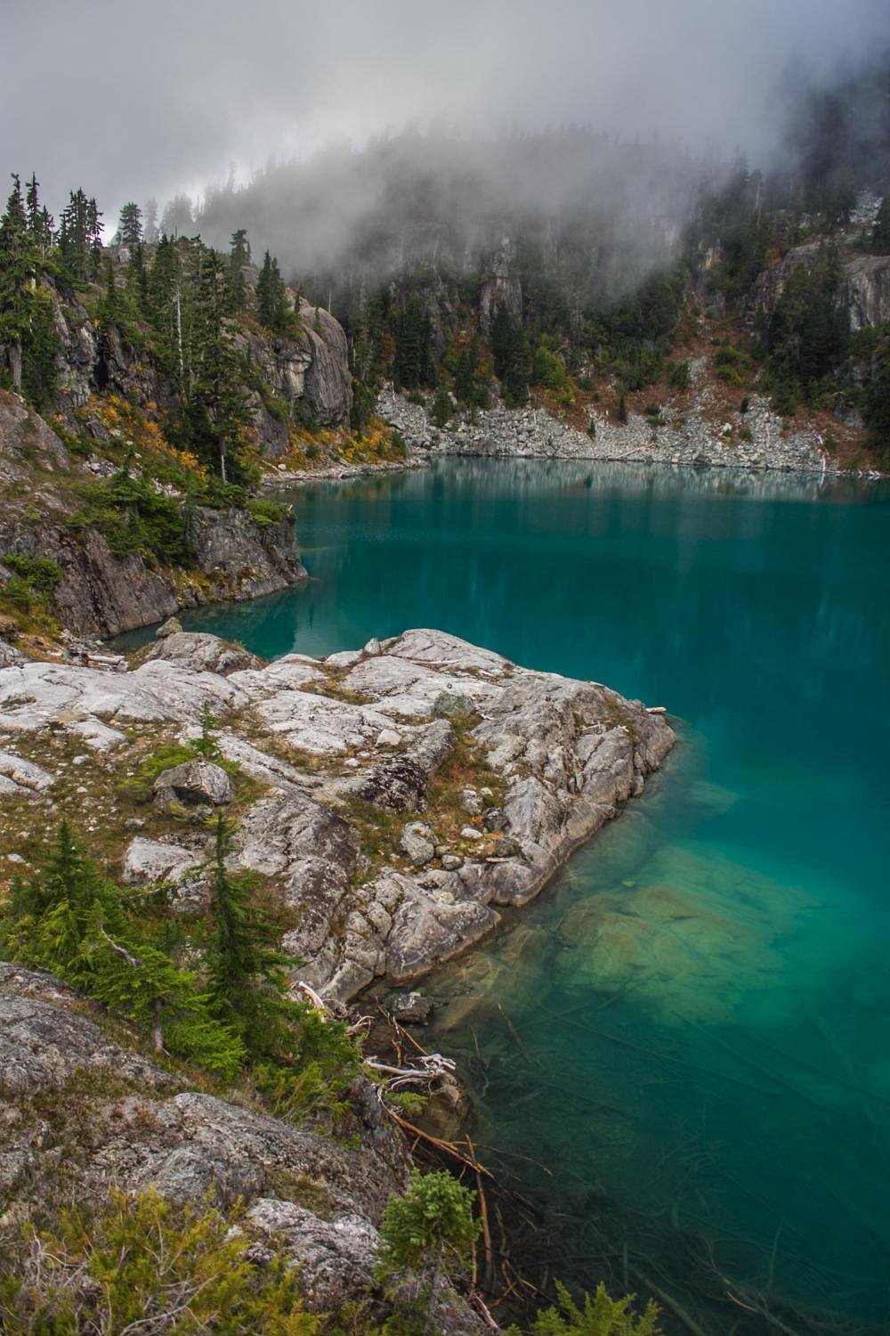 A scenic mountain lake surrounded by rocky cliffs, pine trees, and foggy mountains in the background.