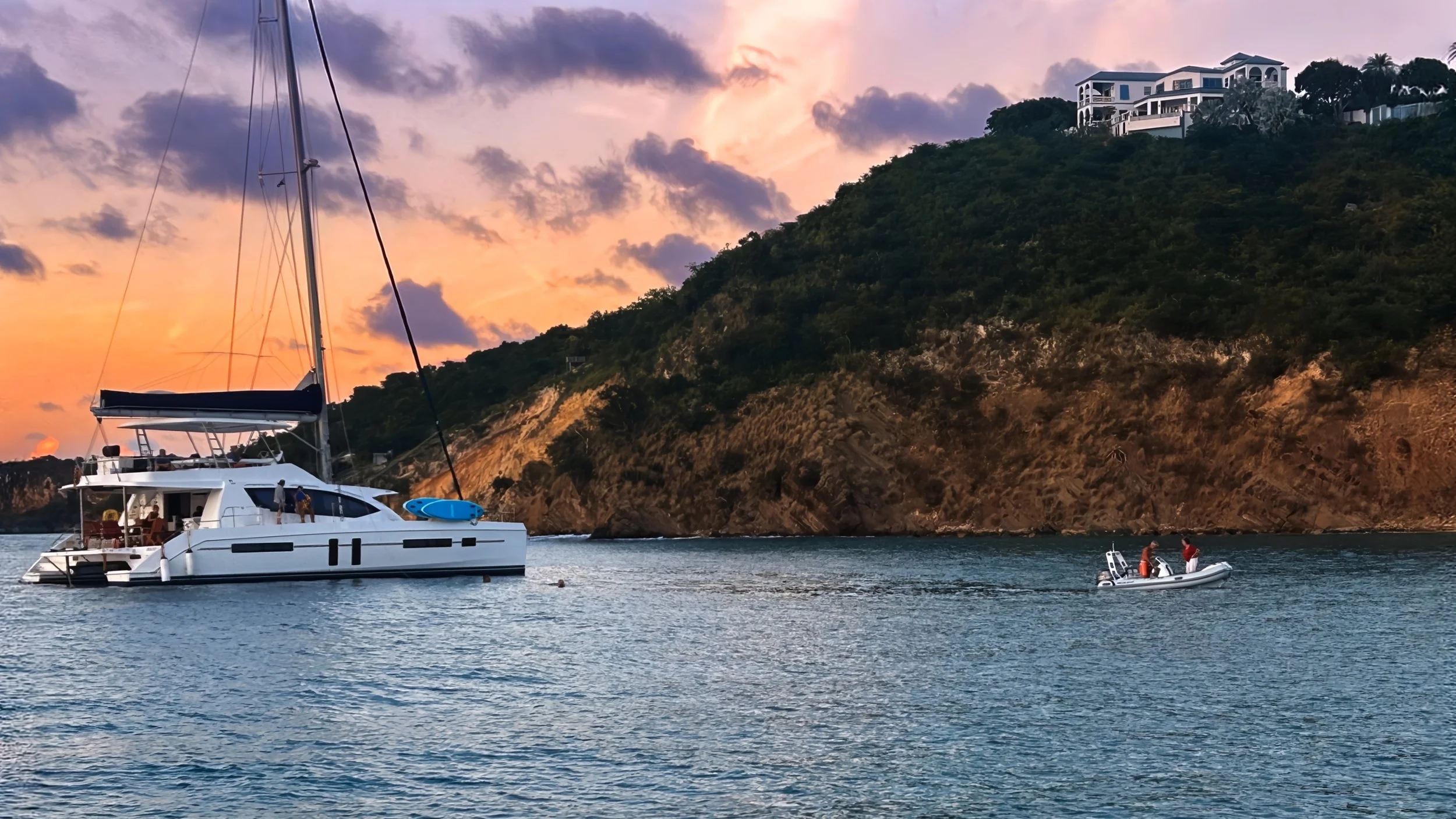 Sailboat and small motorboat on calm water near hillside with house at sunset.