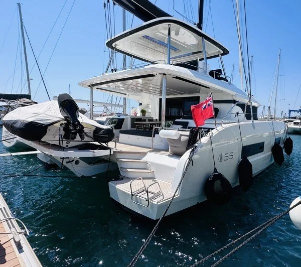 A white luxury yacht docked at a marina, with a small inflatable boat on the stern, and a red flag at the back.