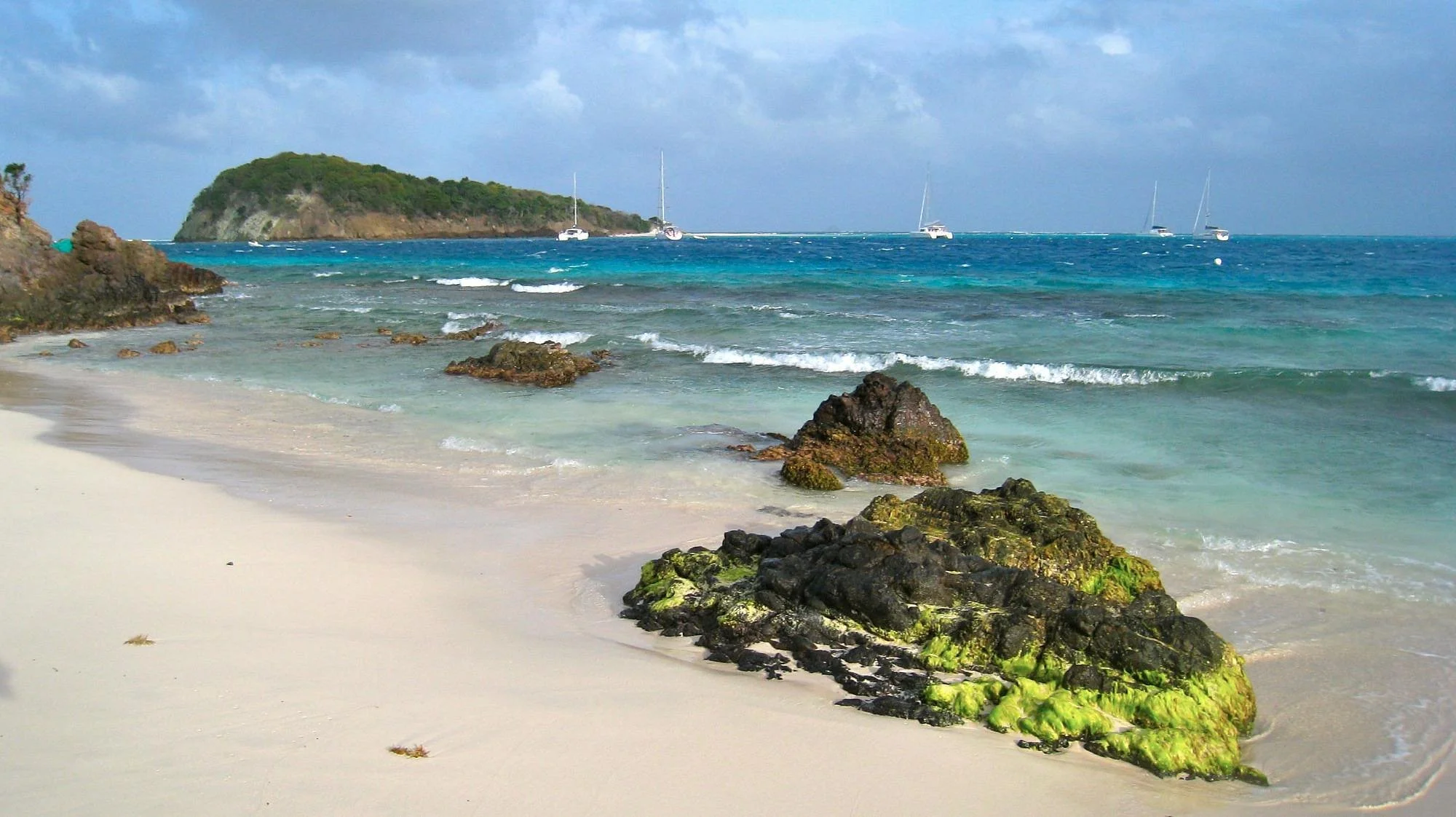 Sunny beach with rocks covered in green moss, clear turquoise water, and sailboats in the distance under a partly cloudy sky.