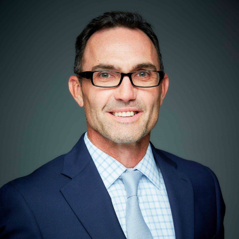 Professional portrait of a man wearing a suit, tie, and glasses, smiling at the camera against a dark gradient background.
