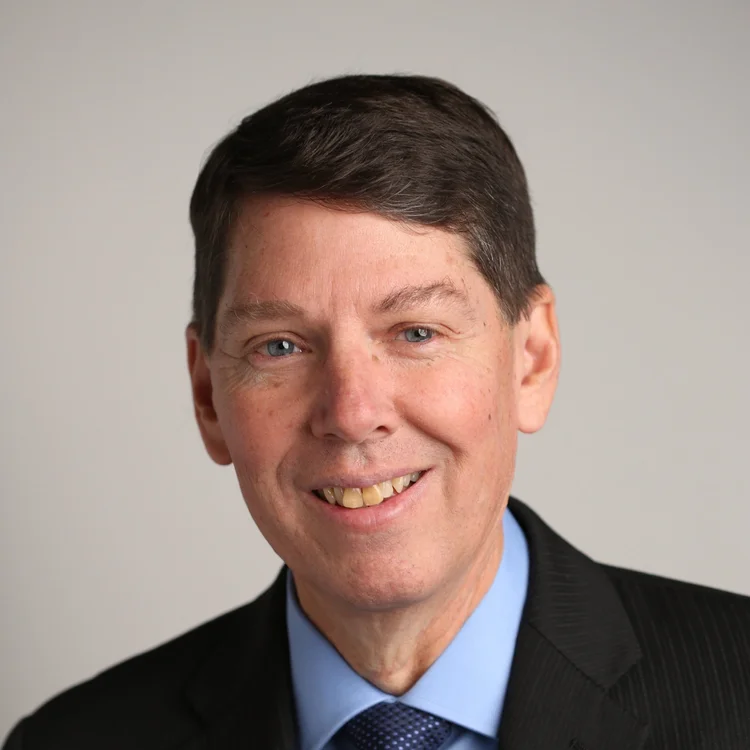 Professional headshot of a middle-aged man with short dark hair, blue eyes, wearing a black suit, blue shirt, and blue tie, smiling against a light gray background.
