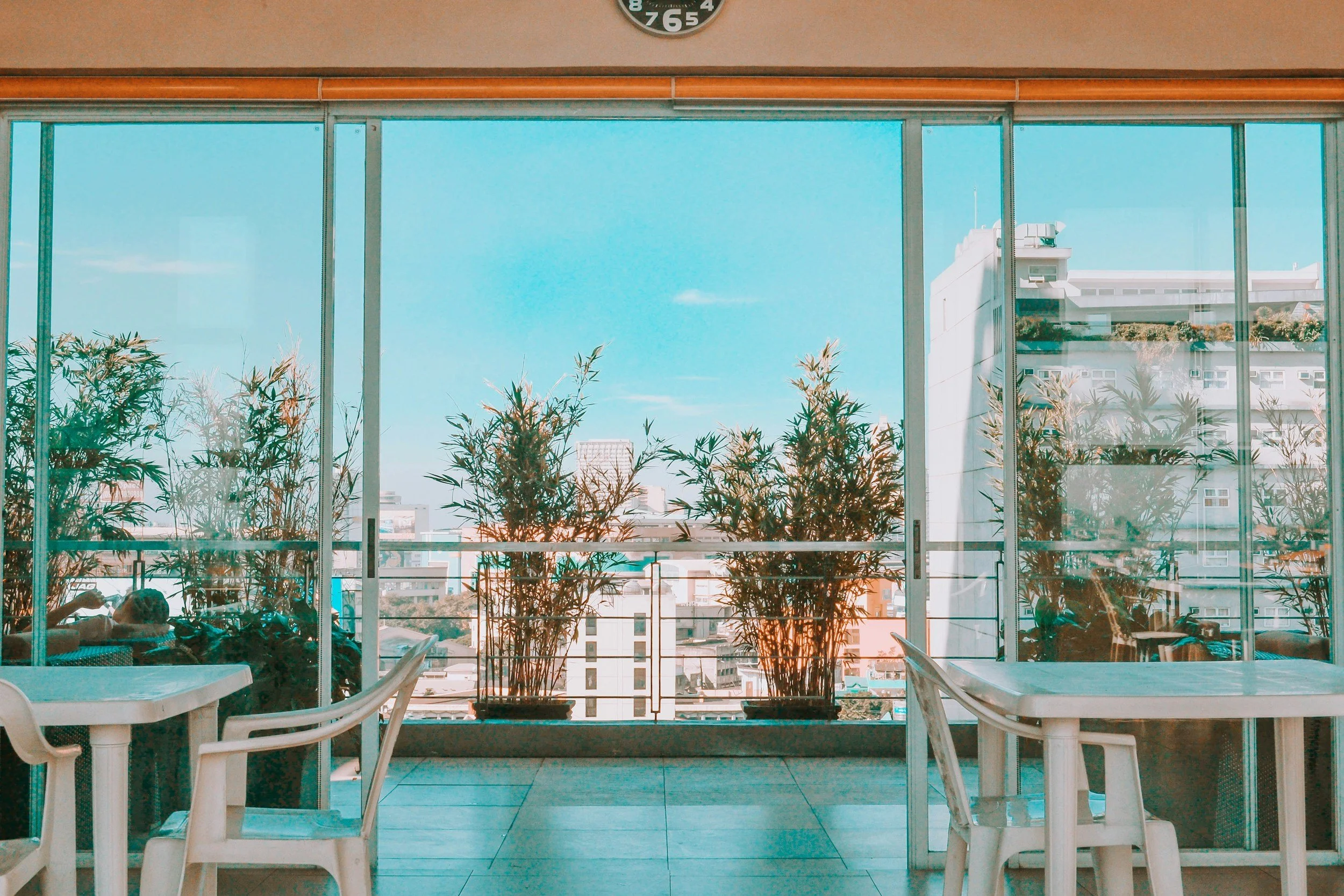 Indoor seating area with white plastic tables and chairs, potted plants, large glass windows showing a cityscape, and a blue sky.