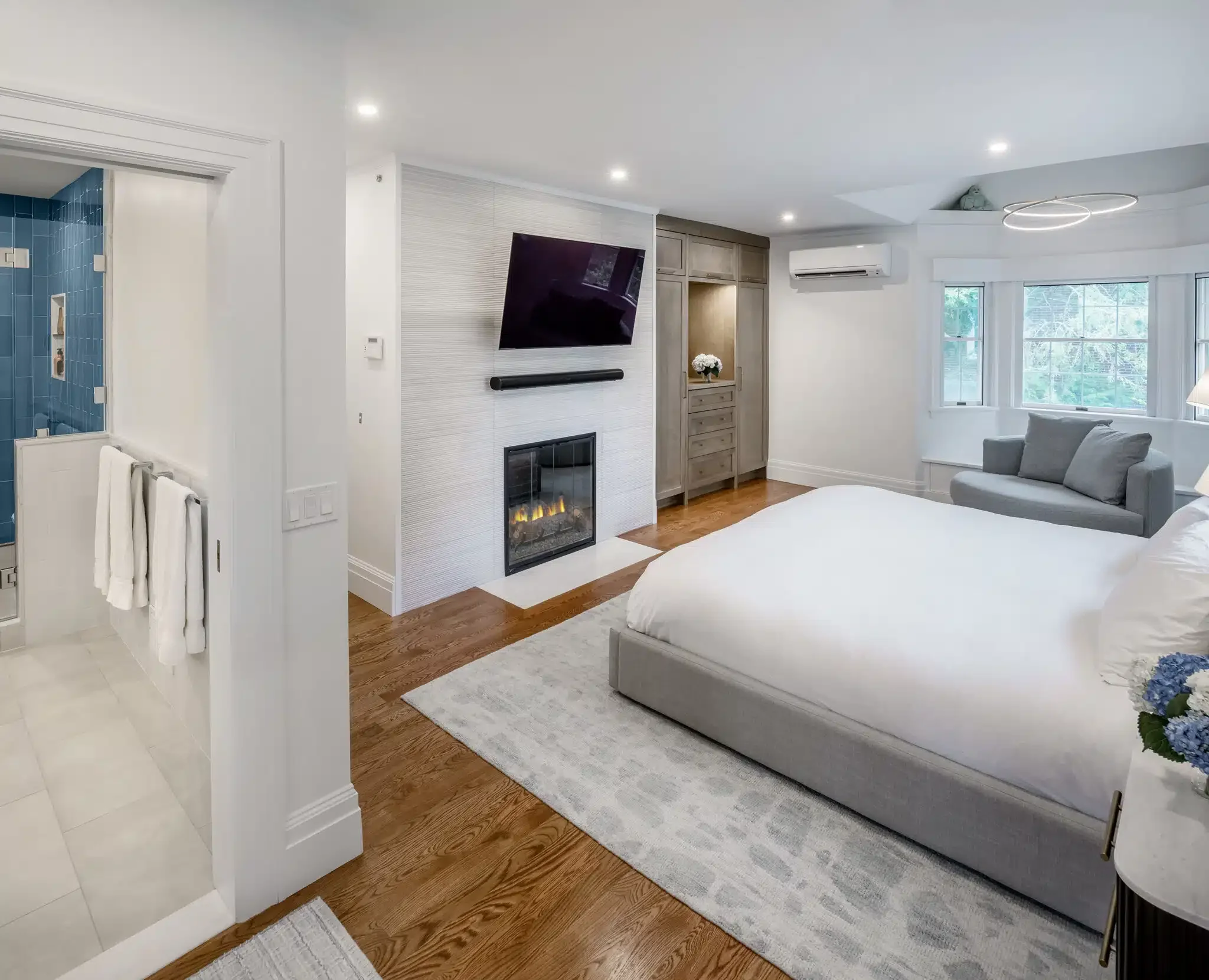 Modern bedroom with a white bed, a gray sofa by a bay window, a wall-mounted flat-screen TV, a fireplace, and wooden flooring.