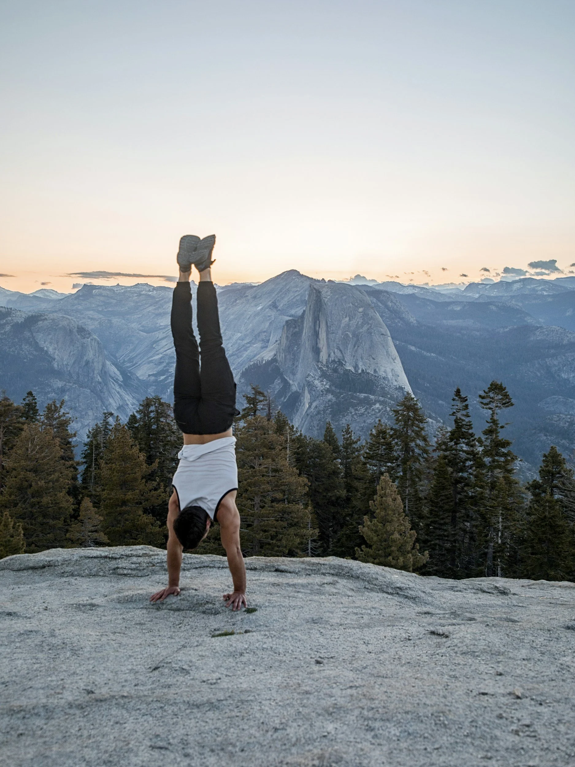 Persona haciendo un handstand en un acantilado con vista a las montañas en el fondo durante el atardecer.