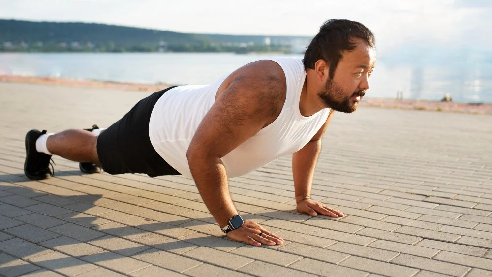 Man doing push-up outdoors on a paved surface near water