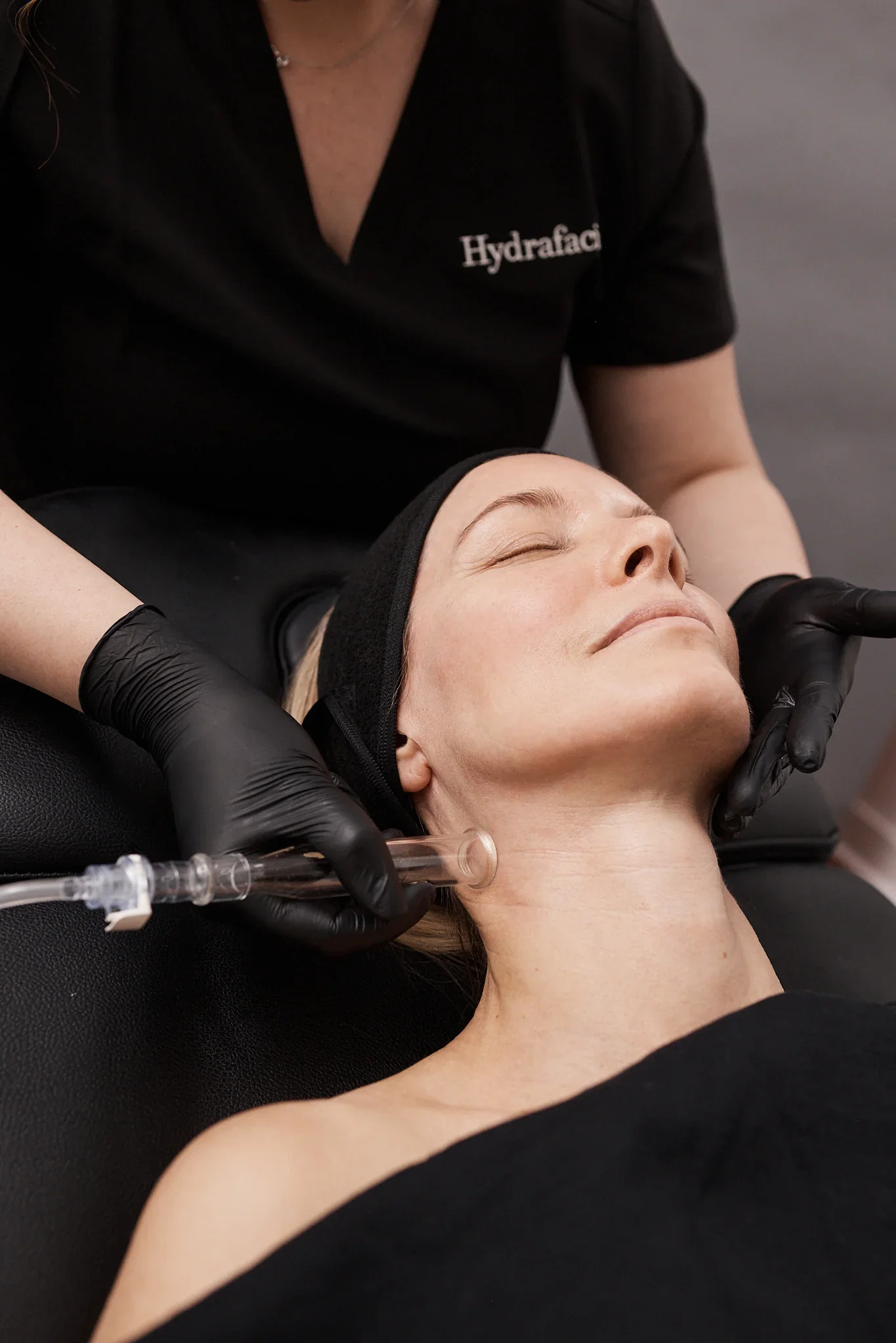 A woman is receiving a facial treatment from a professional in a clinical setting. The professional is wearing a black shirt with the word 'Hydrafacial' embroidered on it. The woman is lying down with her eyes closed, and the technician is using a handheld device on her face, surrounded by a black-gloved hand.