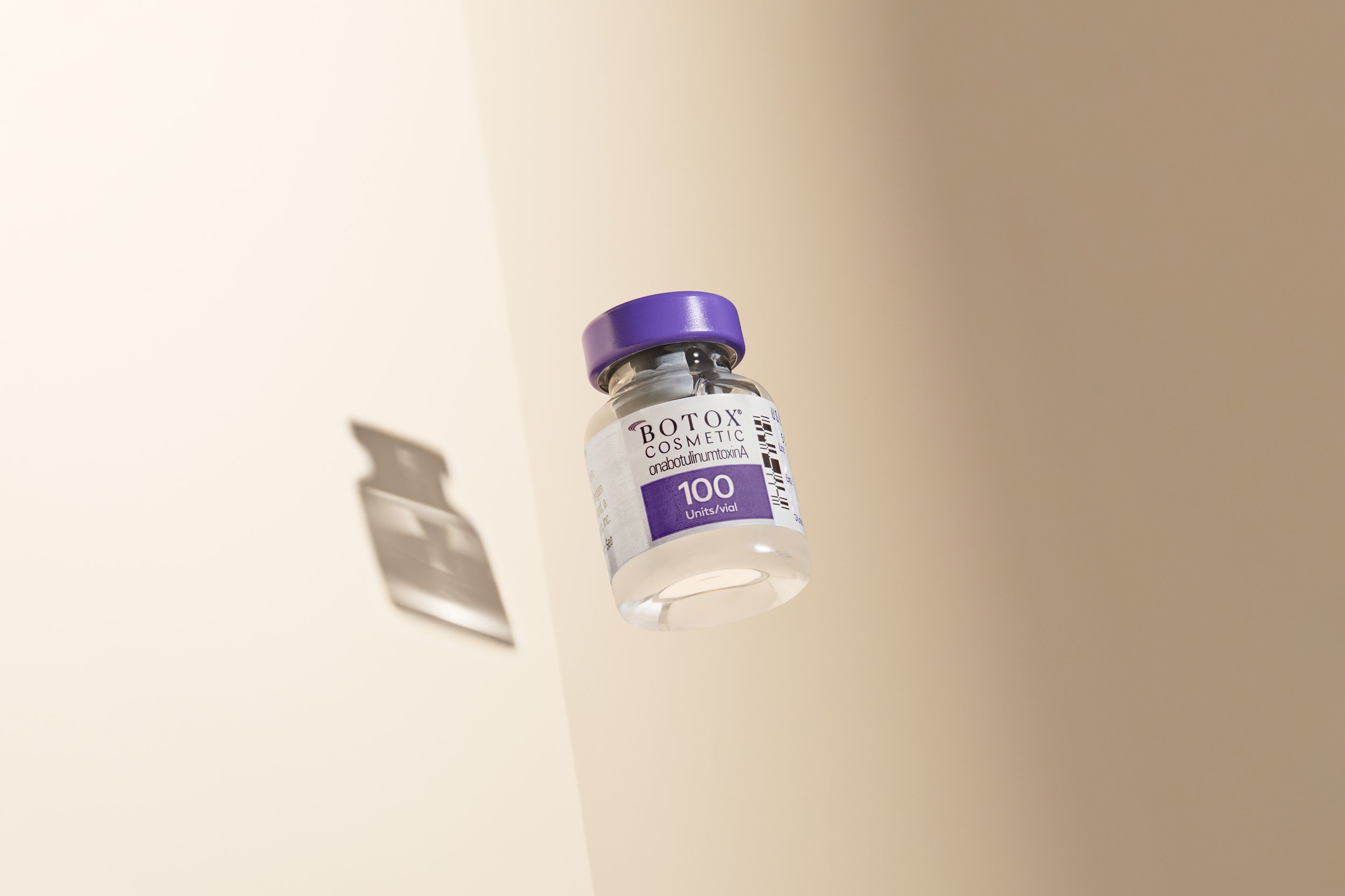 A small glass vial of Botox cosmetic with a purple cap, labeled with '100 units/vial' and 'onabotulinumtoxinA', floating against a divided beige and white background.
