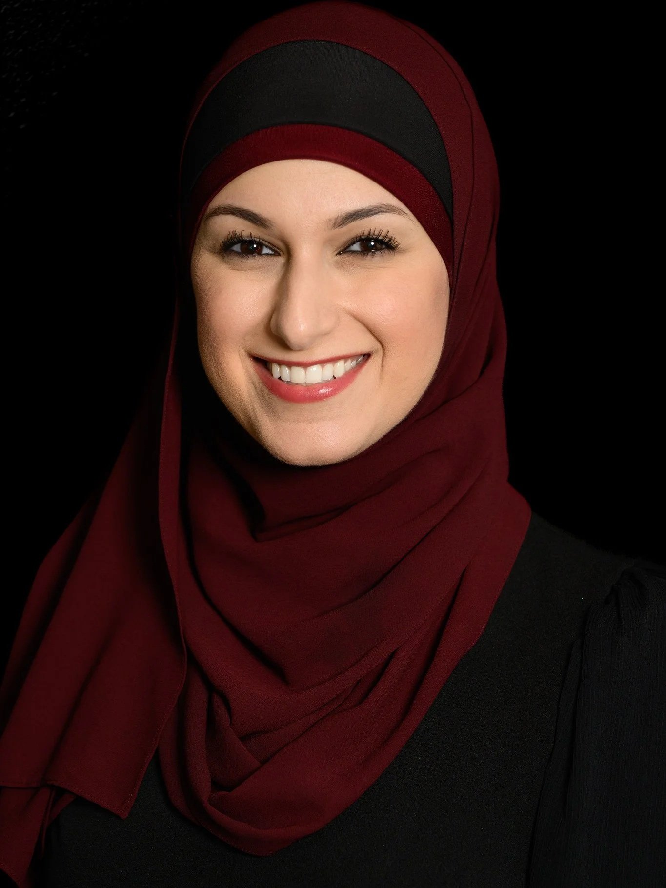 Nurse Practitioner Yusra Awad with a cheerful expression, wearing a burgundy and black hijab and a black top, against a black background.