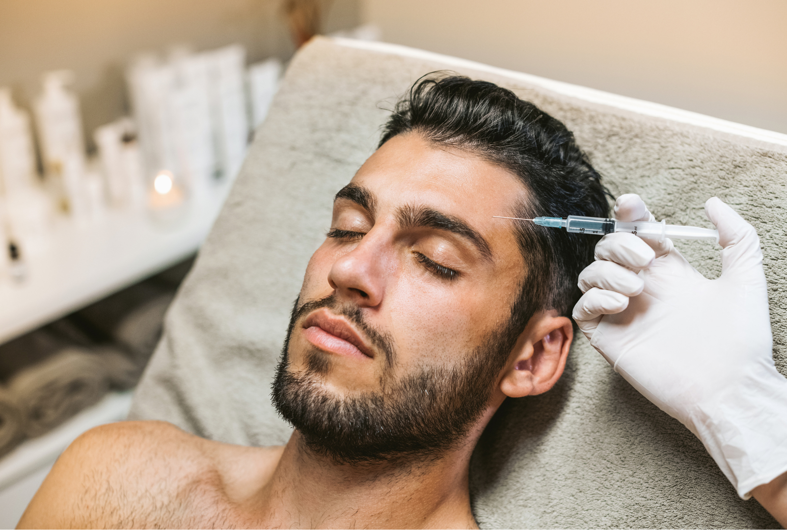 A man with dark hair and a beard receives a cosmetic injection in his forehead from a medical professional wearing white gloves, lying on a bed or examination table.