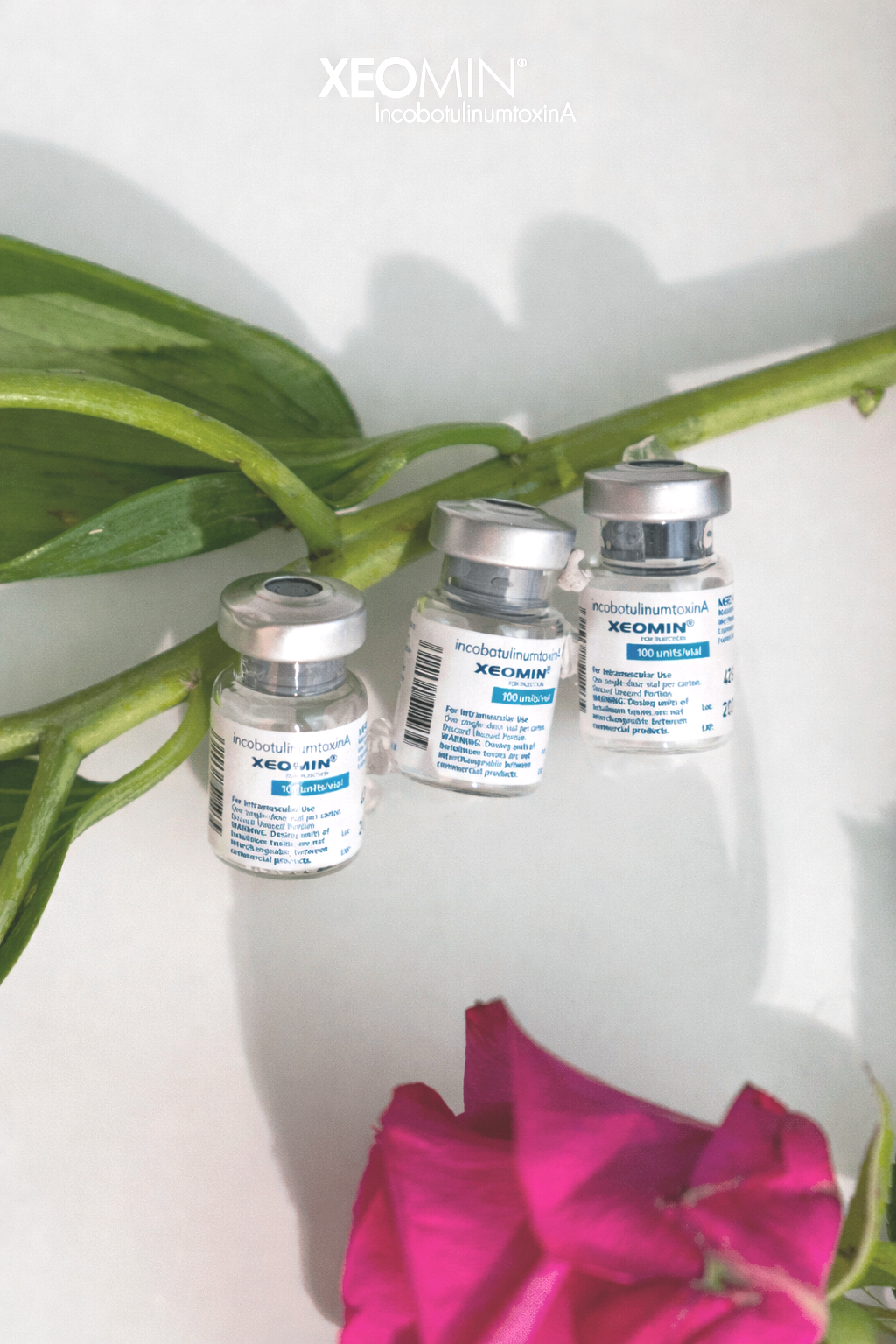 Three vials of XEOMIN, a brand of botulinum toxin A, are placed on a white surface with a green plant stem and leaves behind them and a pink flower at the bottom of the image.