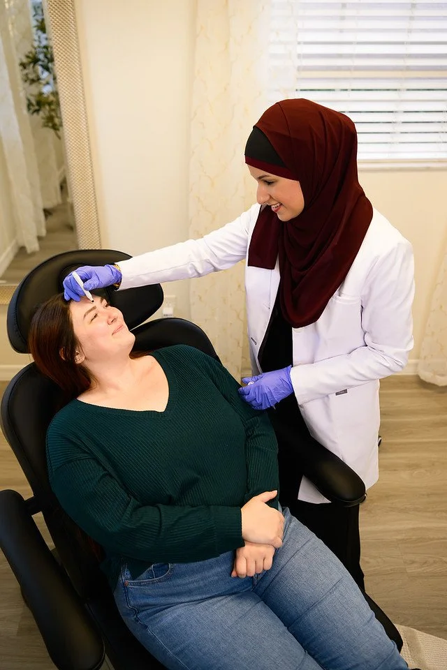 Nurse Practitioner Yusra Awad is giving a Botox injection to a woman lying back in a chair, who appears relaxed and smiling.
