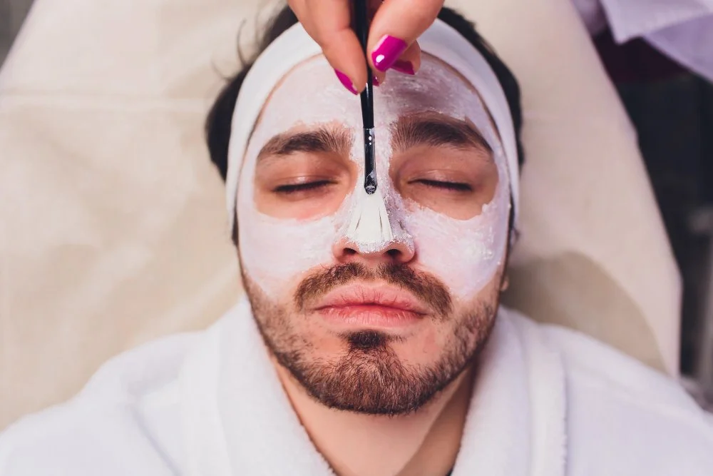 A man with a beard and closed eyes receives a facial treatment, with a white facial mask applied, while a person with pink nail polish uses a brush to apply or remove product on his face.
