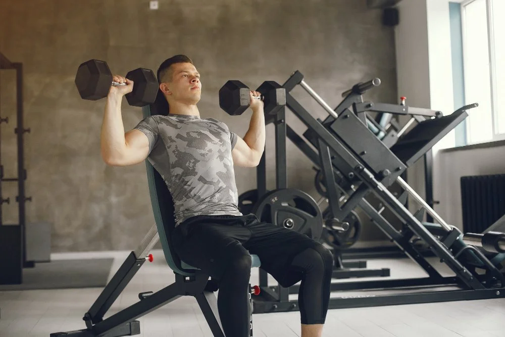 A young man in gray camouflage t-shirt and black leggings sitting on a gym bench, lifting two black dumbbells in a gym with exercise machines and a window in the background.