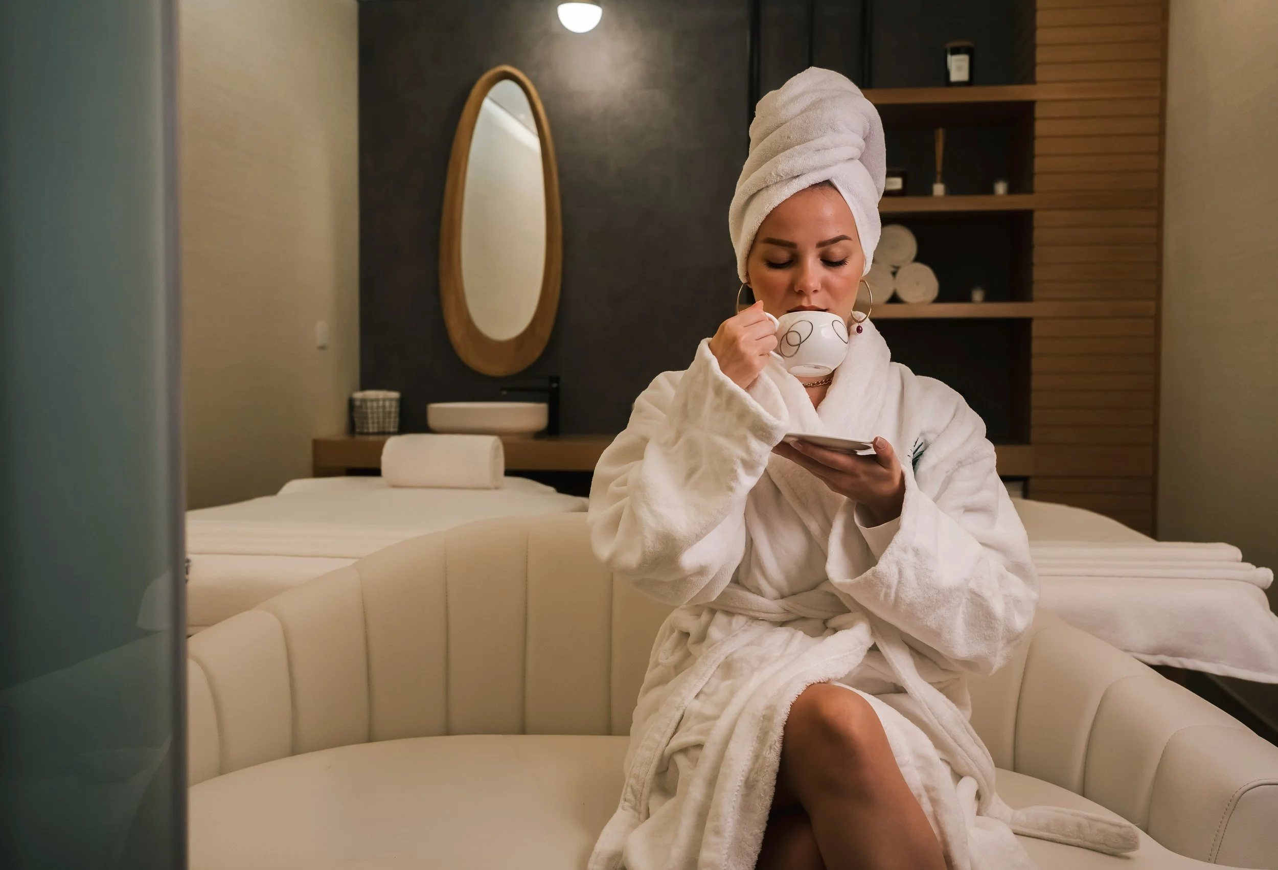 Woman in a white bathrobe with a towel wrapped around her head, sitting on a cream-colored curved sofa, drinking from a cup and looking at her phone in a spa or massage room.