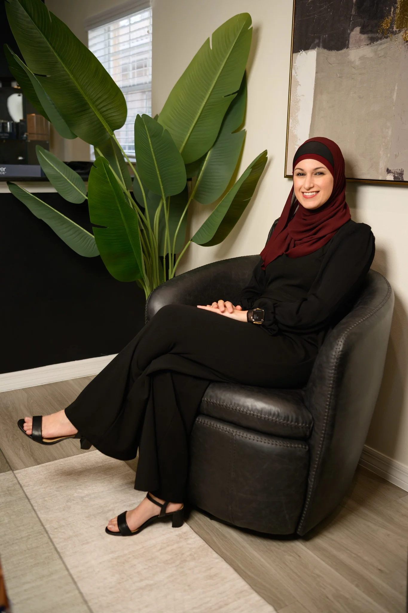 Nurse Practitioner Yusra Awad sitting on a black leather armchair in a living room, smiling, wearing a black outfit, a maroon hijab, and open-toe heels, with a large green plant and an abstract painting on the wall behind her.