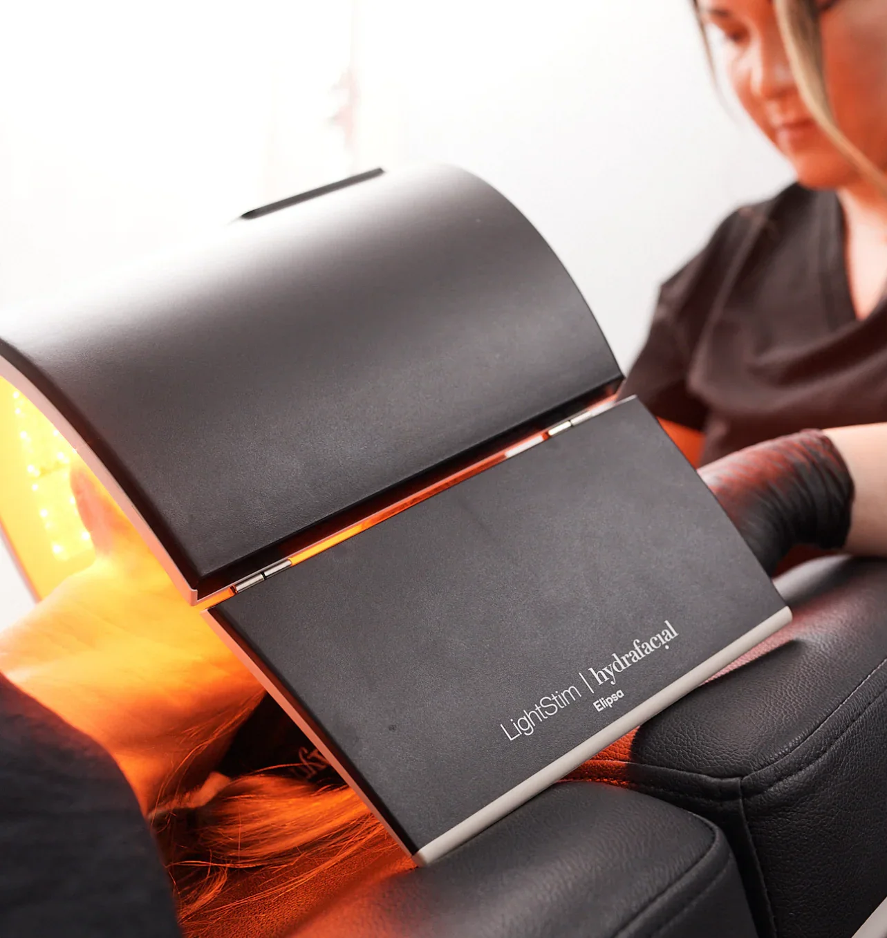 A person lying on a massage table receives a LightStim HydraFacial treatment, with a black laser device emitting orange light.