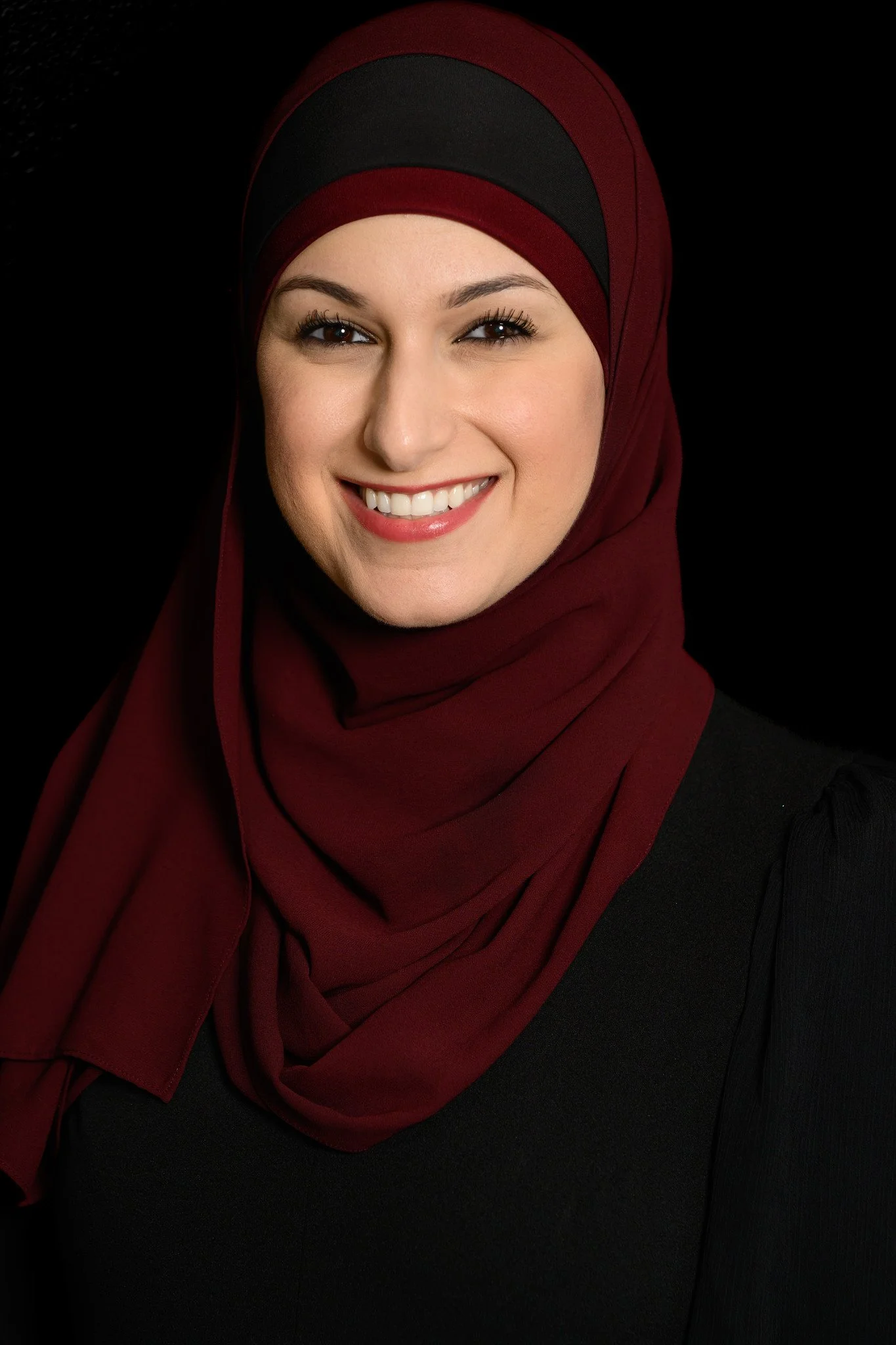 Nurse Practitioner Yusra Awad with a cheerful expression, wearing a burgundy and black hijab and a black top, against a black background.