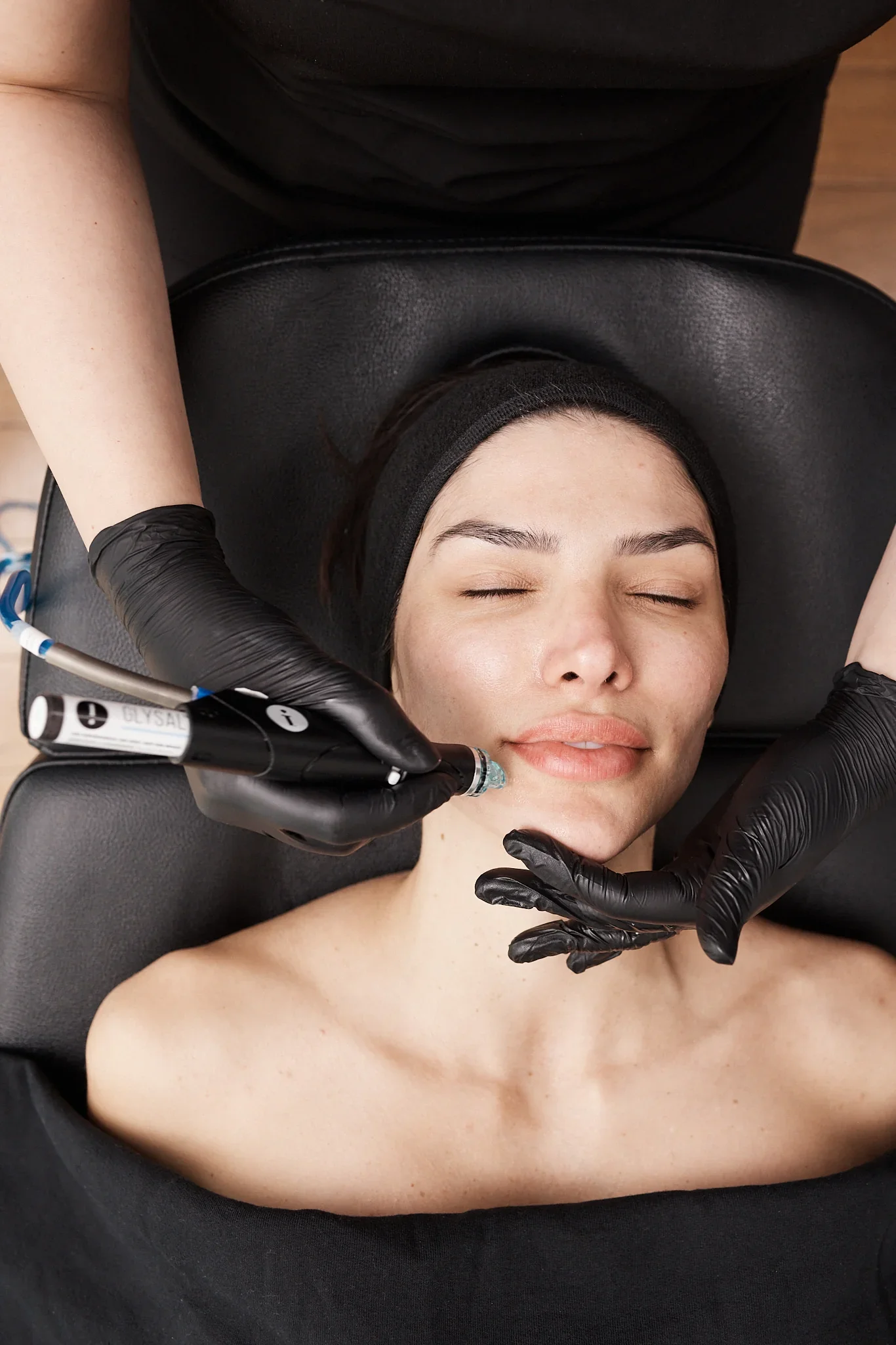 A woman with closed eyes receives a cosmetic injection in her face, lying on a black treatment bed with a black headband, while a healthcare professional in black gloves administers the injection.