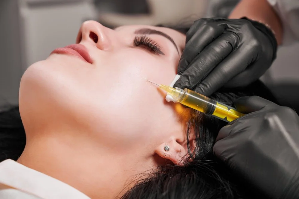 Person receiving a cosmetic treatment, lying down, with a medical professional injecting a syringe into her face.