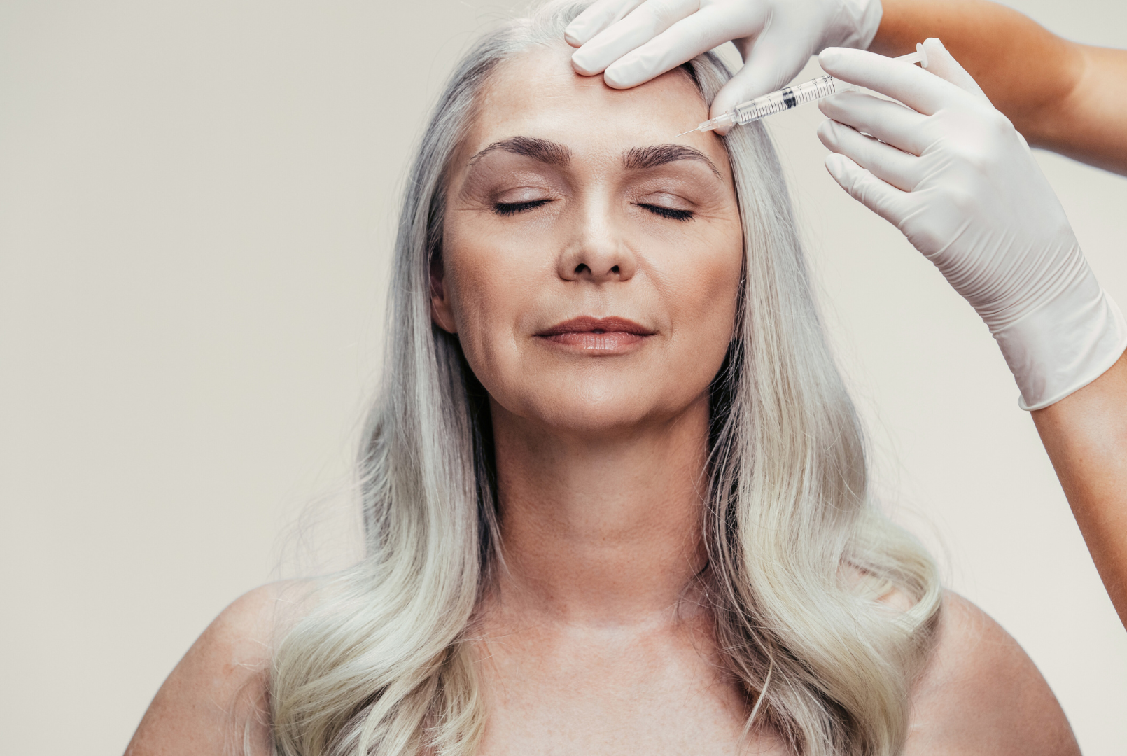 A woman with gray hair relaxing with her eyes closed as a medical professional administers an injection to her forehead.