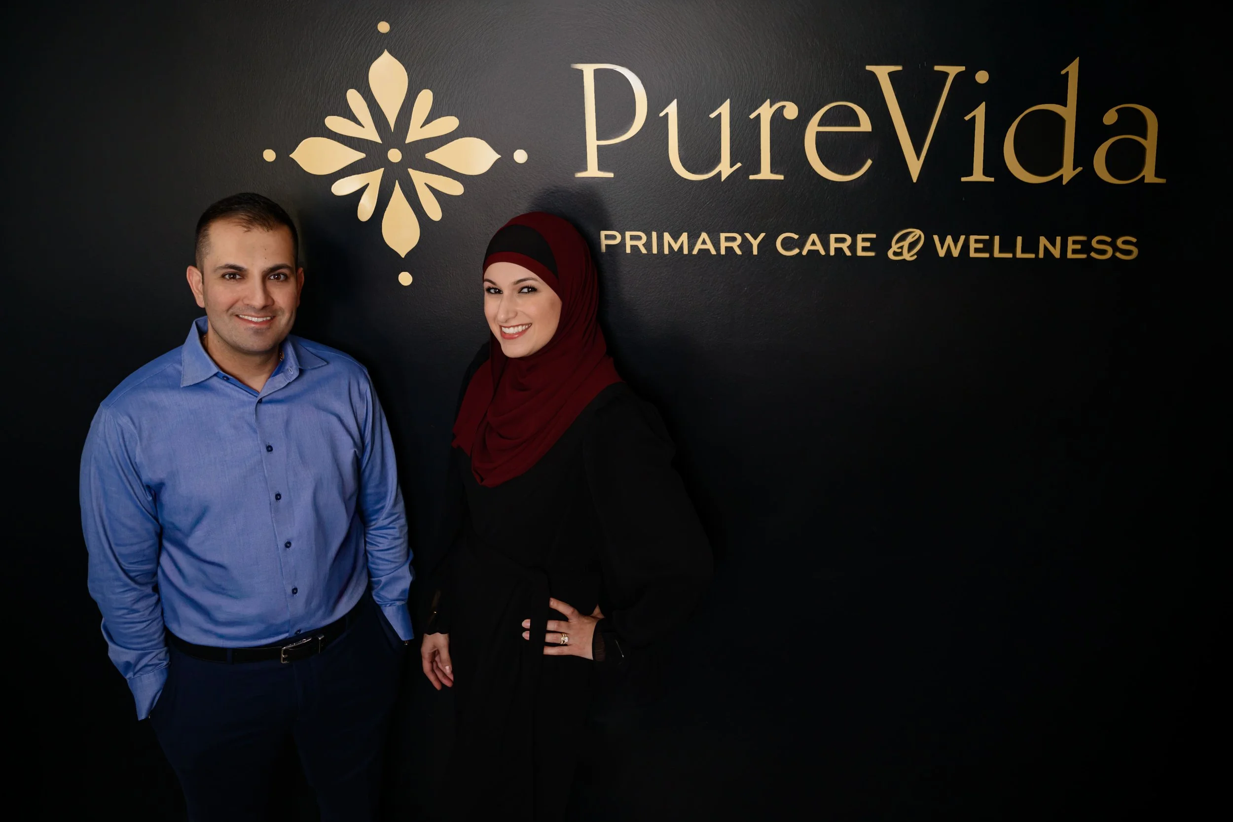 Dr. Said Awad Nurse Practitioner Yusra Awad standing in front of a black wall with a gold logo and text that says PureVida Primary Care & Wellness.