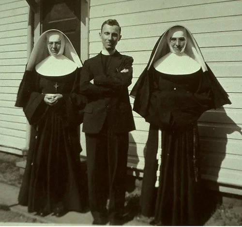 A man standing between two women dressed as nuns in front of a wooden building.