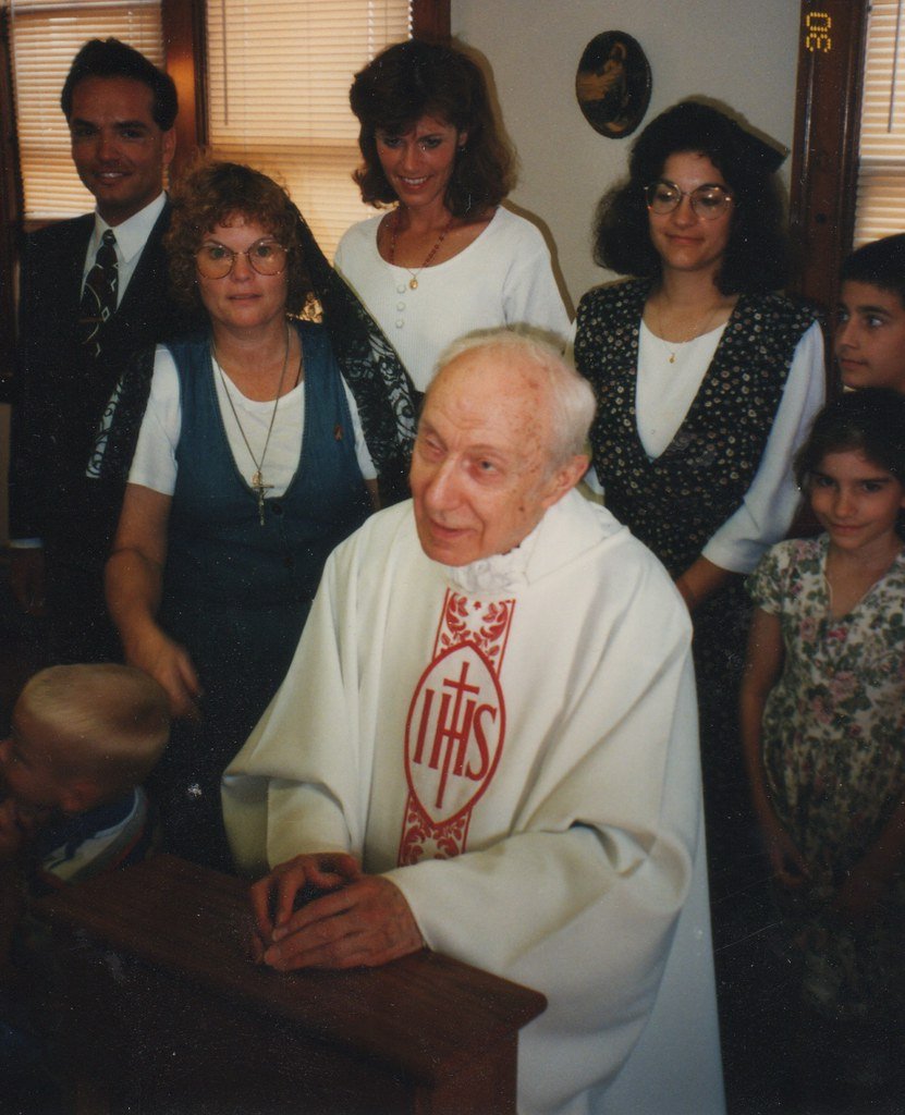 An elderly priest in white vestments with red embroidery and a cross, sitting at a table in a room with seven people standing behind him, including women, children, and a man, all smiling during a gathering or celebration.