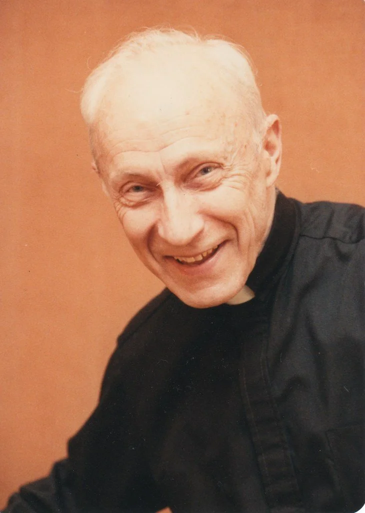 A smiling elderly man with white hair, wearing a black clerical shirt with a white collar, against a warm beige background.