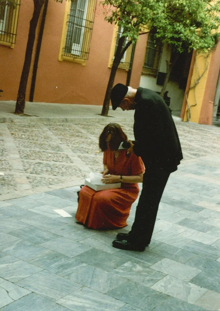 A woman in a red dress sitting on a bench receives a gift from a man in a black suit and hat on a city sidewalk with colorful buildings and trees.