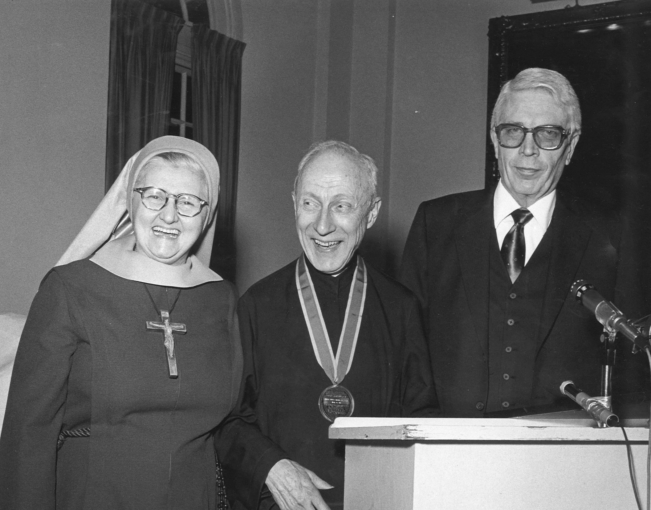 A nun, a man with a medal, and a man with glasses at a podium, smiling and standing together during a ceremony.