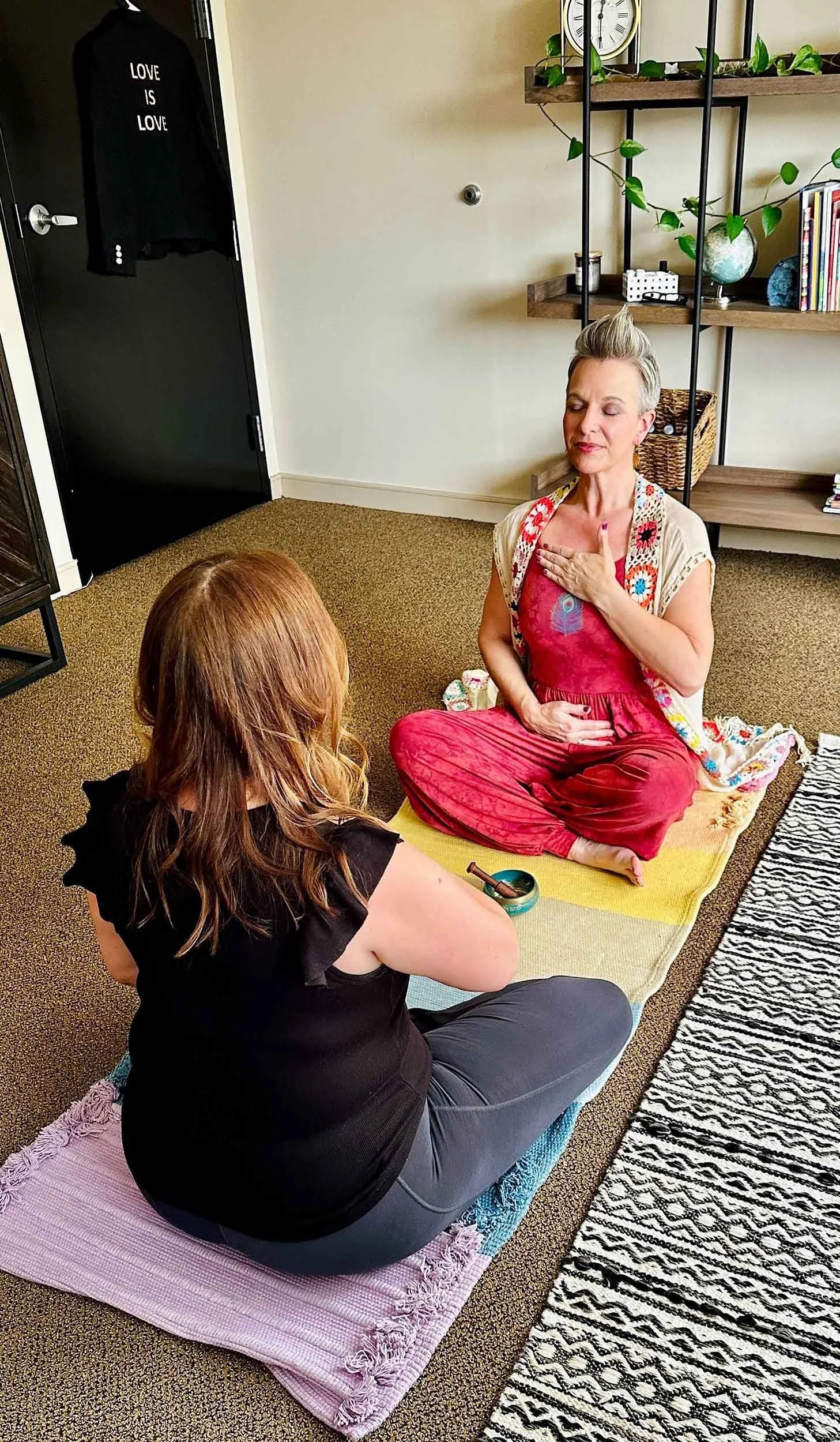 Two women are sitting on yoga mats in a room, one with red hair and black top, the other with gray hair and colorful clothes. The gray-haired woman has her eyes closed, one hand over her heart, and the other on her stomach, practicing meditation or relaxation.