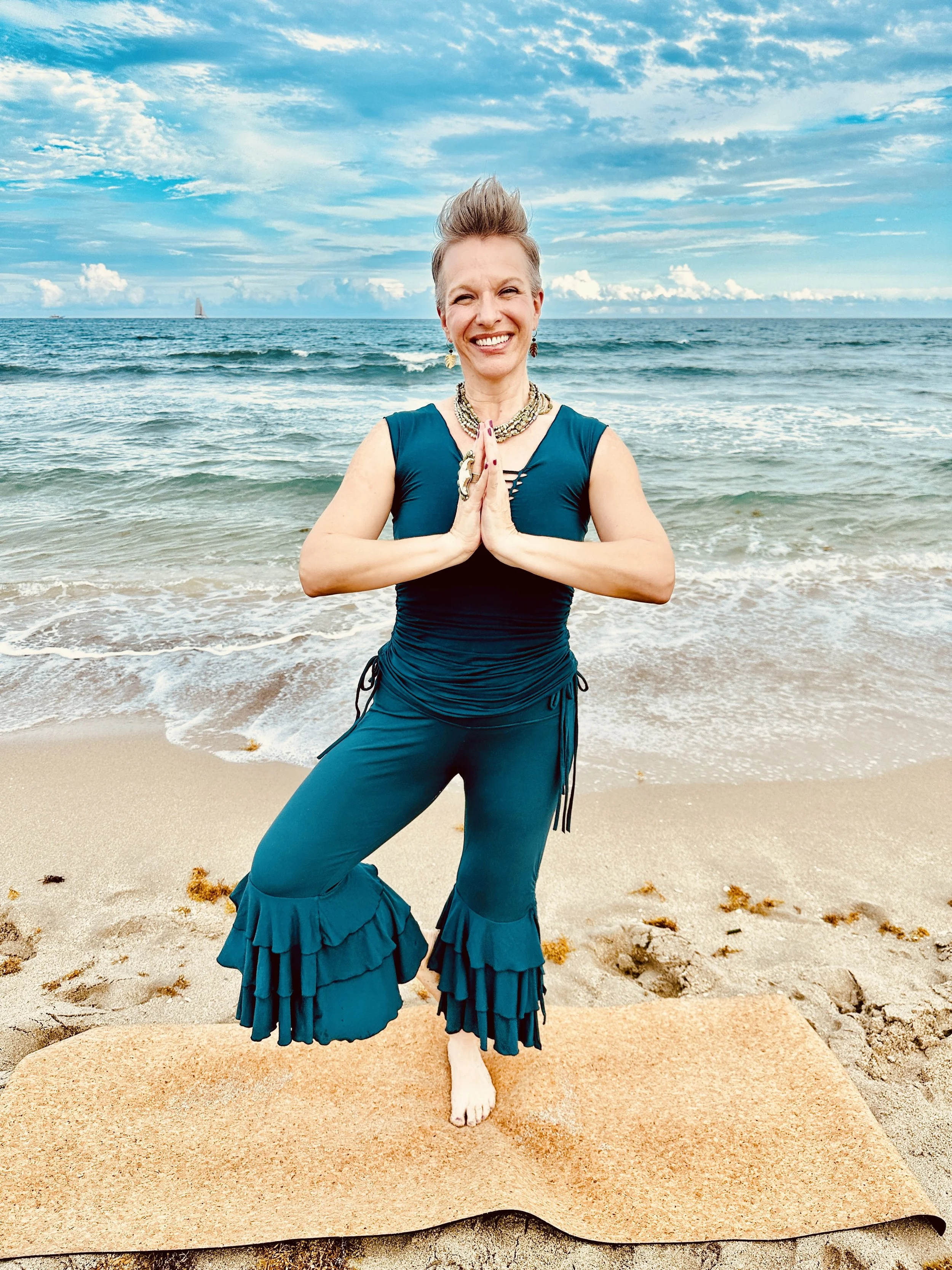 Angela Towne practicing yoga on a beach, standing on one foot with hands in prayer position, smiling, with ocean waves and a cloudy blue sky in the background.