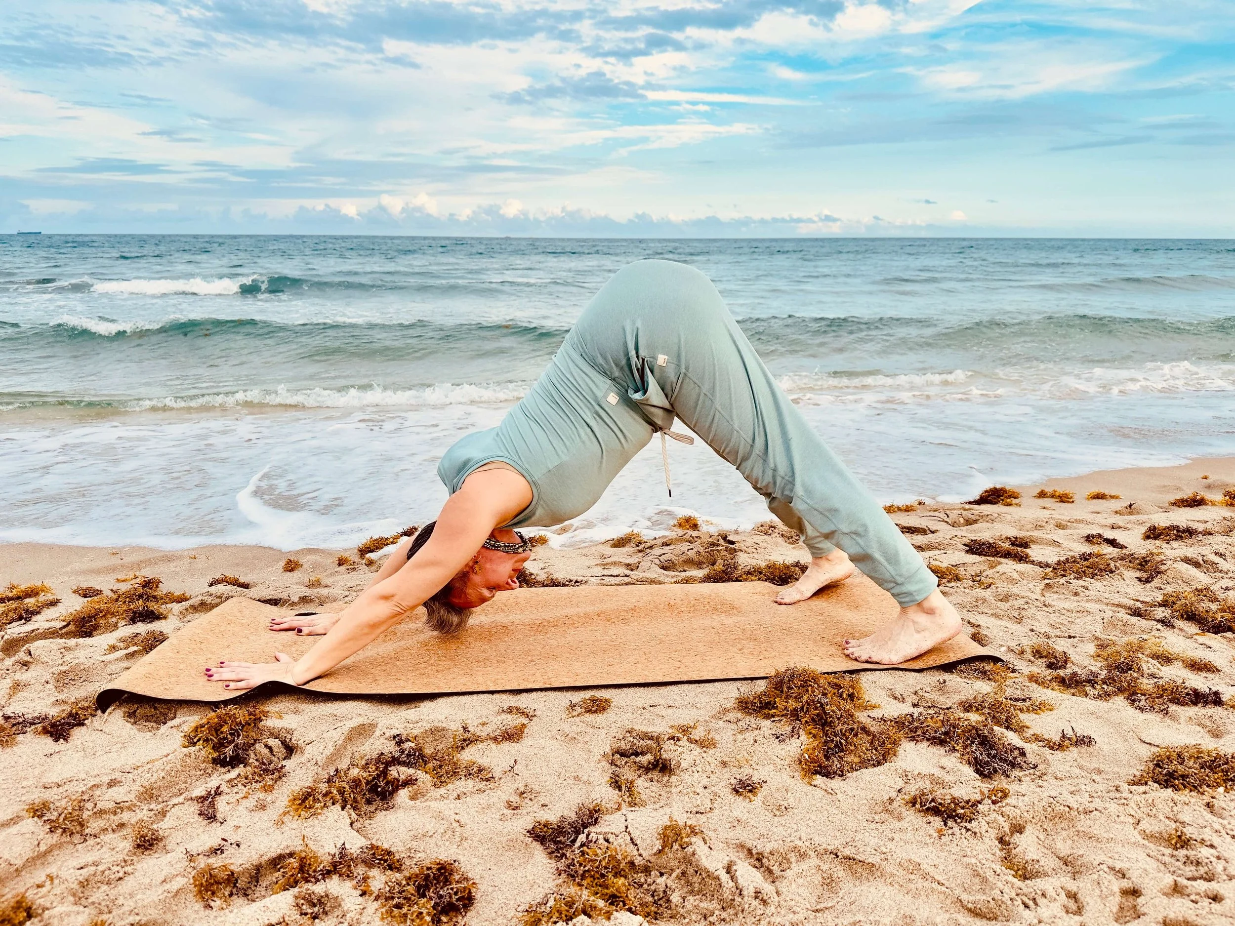 Woman practicing yoga on a beach, performing a downward dog pose with the ocean and sky in the background.